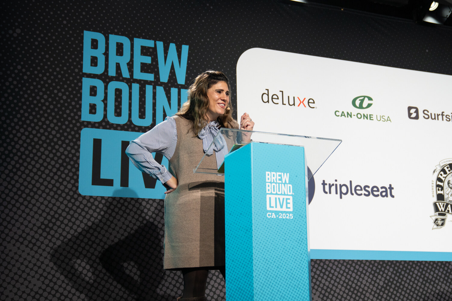 Woman in a tan vest and striped blouse standing at a Brewbound Live podium in front of a sponsor wall.