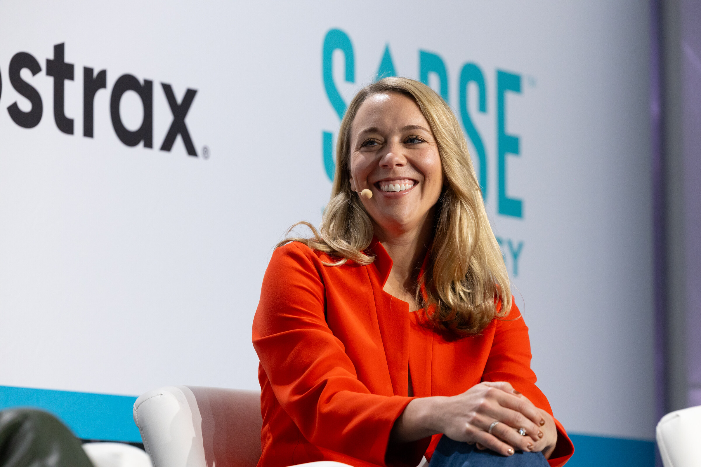Smiling woman in an orange blazer seated on stage in front of Abstrax and Sorse Technology logos.