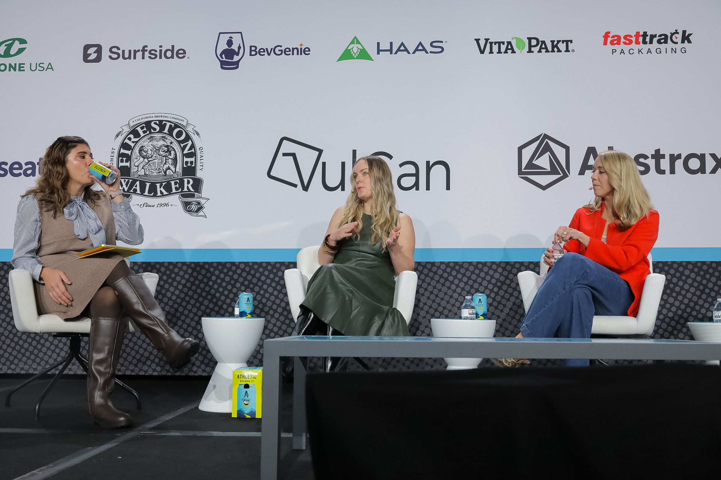 Three women in conversation on white chairs, one drinking from a can, in front of a Firestone Walker logo.