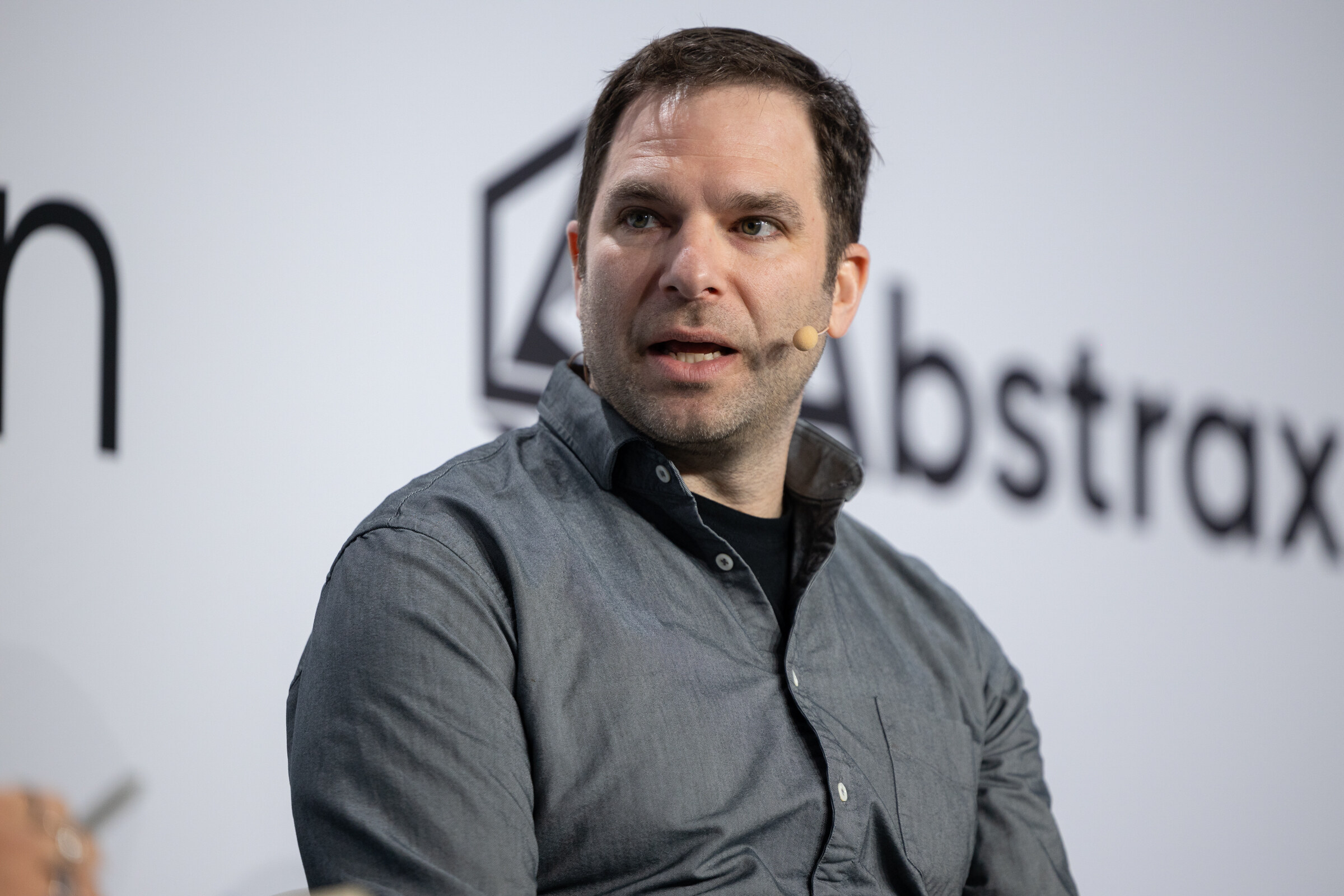 Man in a gray button-up shirt wearing a headset microphone and speaking in front of an Abstrax logo.