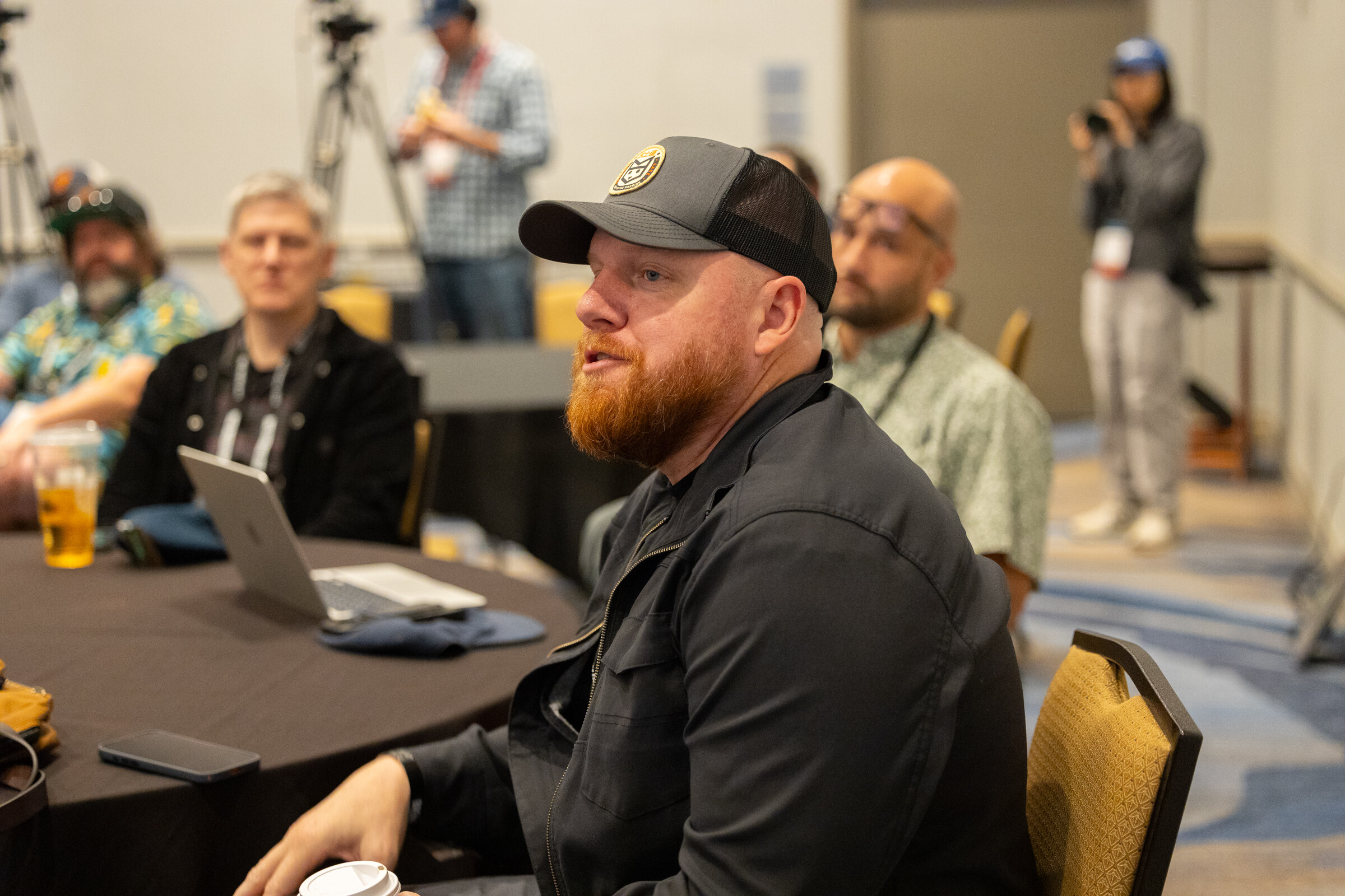 Bearded man in a trucker cap and dark jacket speaking while seated at a round table in a meeting room.