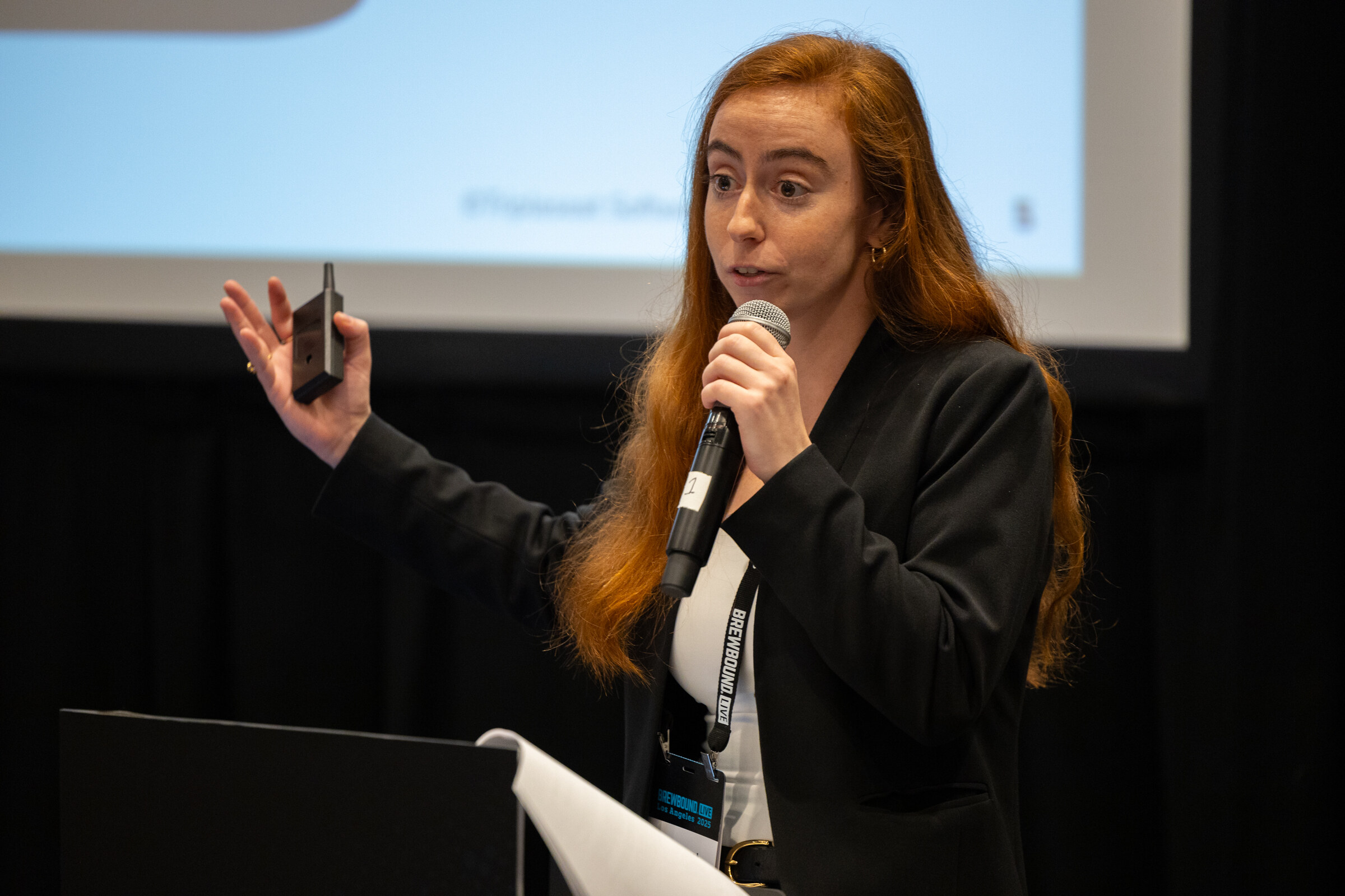 Woman with long red hair holding a microphone and clicker beside a podium and laptop.