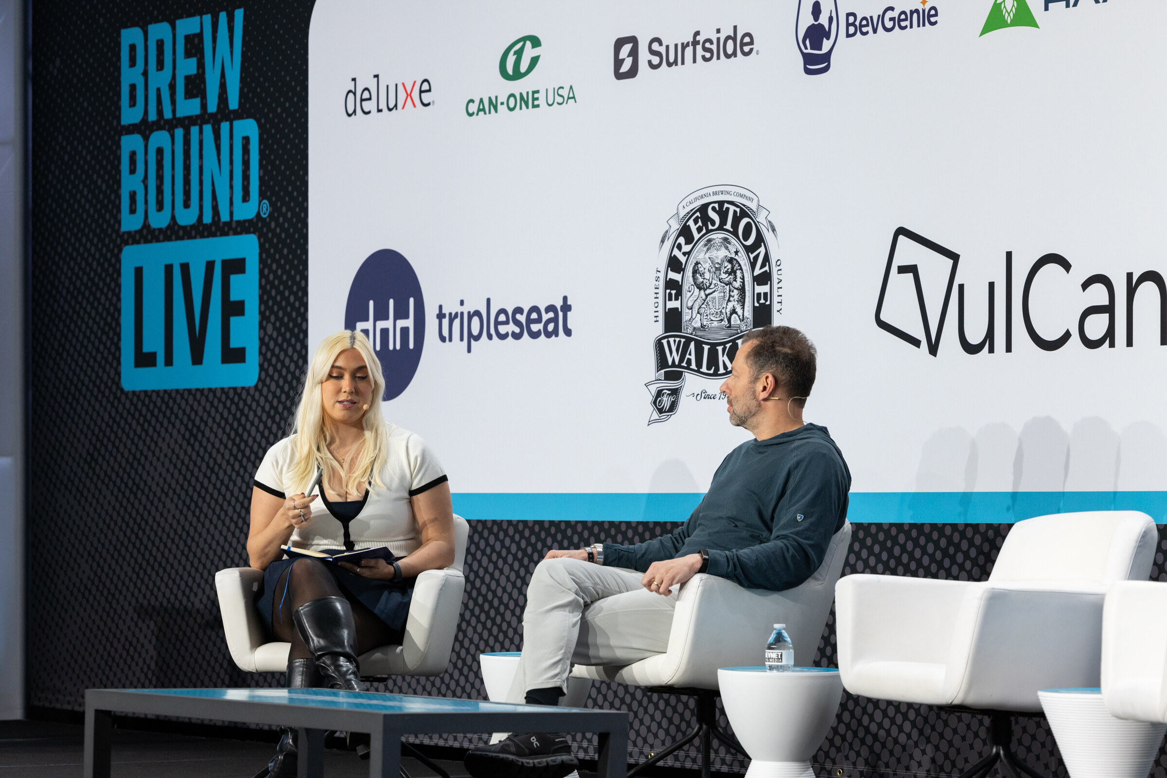 A woman in a white top with a notebook and a man in a hoodie seated on white chairs in front of Tripleseat, Firestone Walker and VulCan logos.