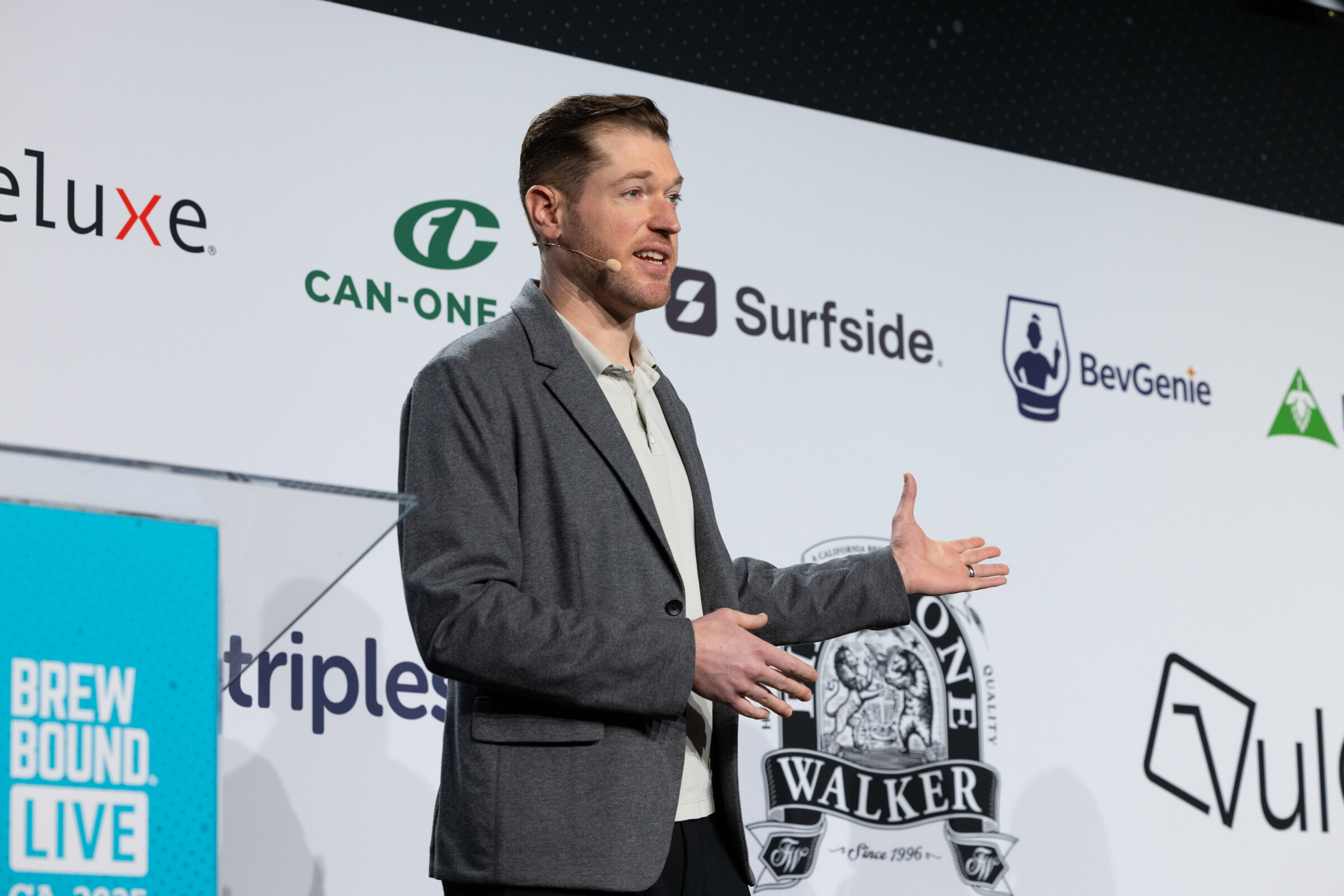 Man in a gray blazer speaking at a podium in front of Deluxe, Can-One USA, Surfside and BevGenie logos.