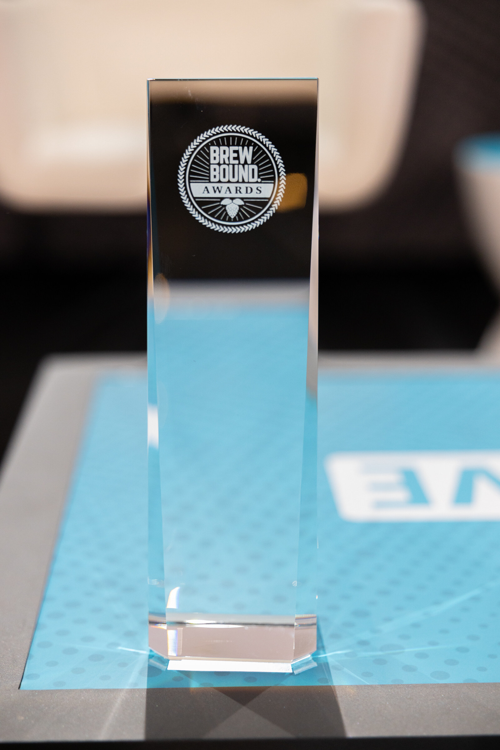 Tall glass award engraved with the Brewbound Awards logo standing on a blue table cover.