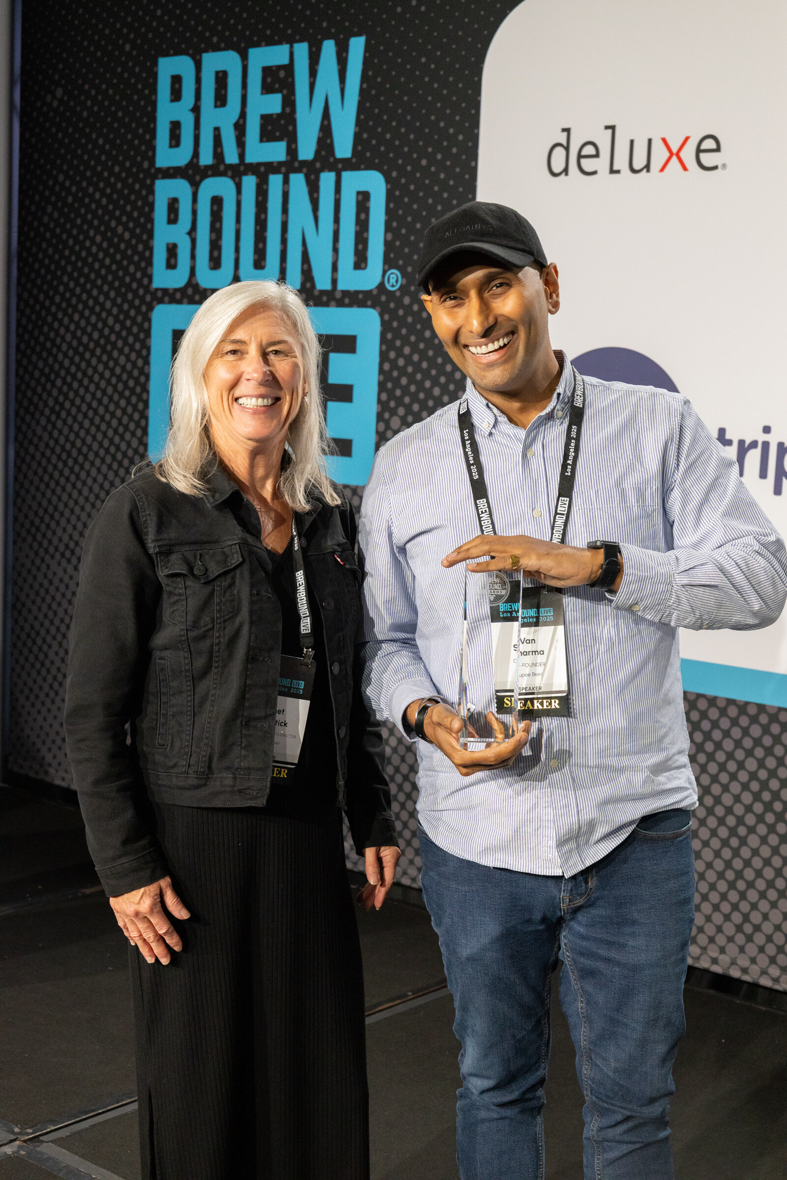 Woman in a black denim jacket and man in a light blue shirt and cap holding a glass trophy in front of a Brewbound Live backdrop.