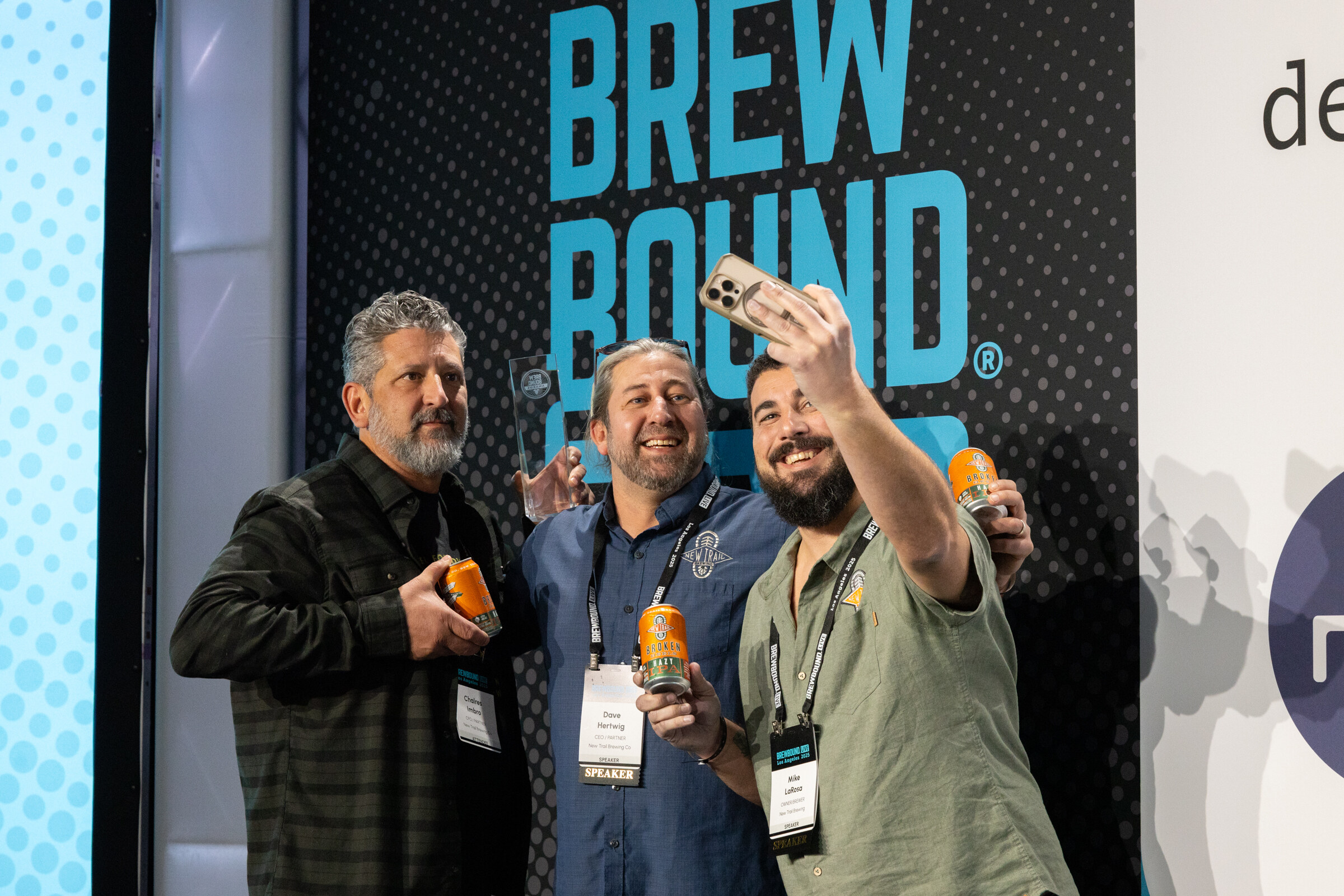 Three men with conference badges holding orange cans in front of a Brewbound Live backdrop while one takes a phone selfie.