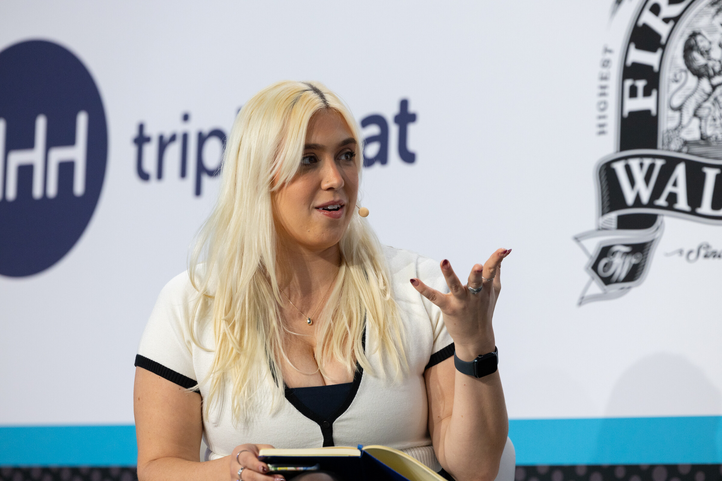 Woman with long blond hair in a white and black top holding a notebook and speaking, in front of Tripleseat and Firestone Walker logos.
