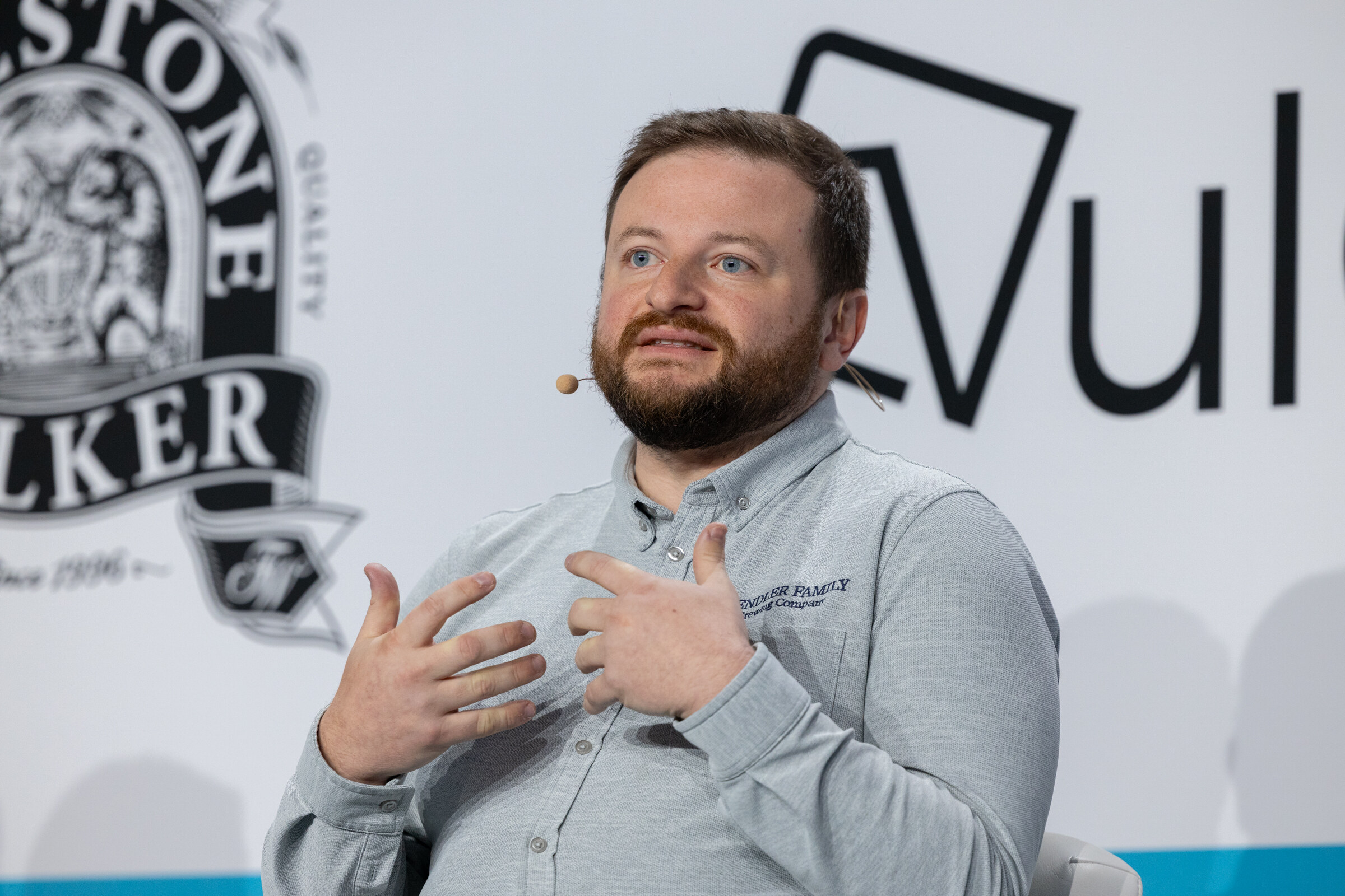 Bearded man in a gray polo with a headset microphone gesturing with both hands in front of a Firestone Walker logo.