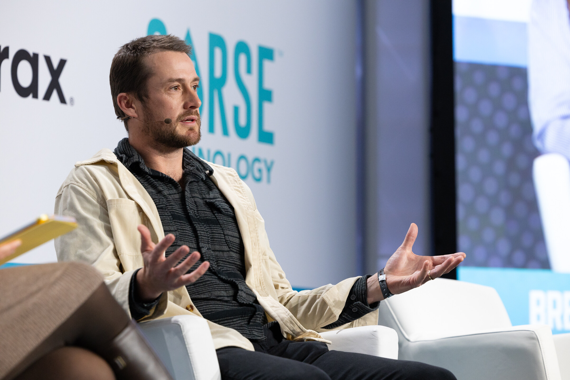 Man in a cream jacket and dark shirt gesturing with open hands while seated on a white chair in front of a Sorse Technology logo.