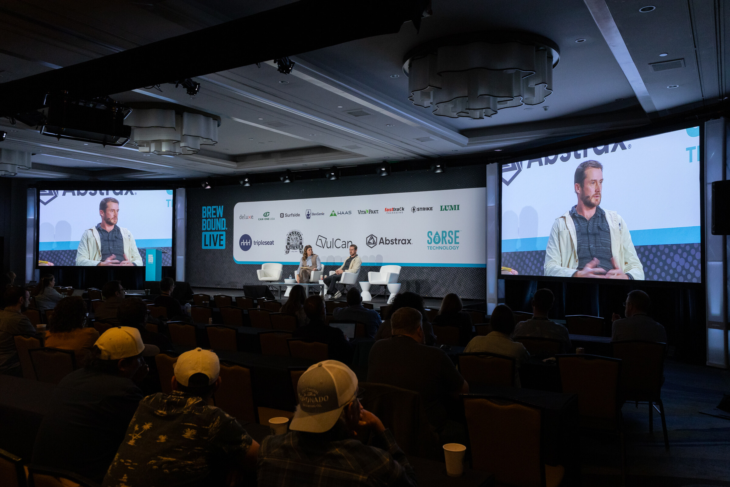 Wide view of two people on a stage with large video screens either side and rows of attendees in the foreground.