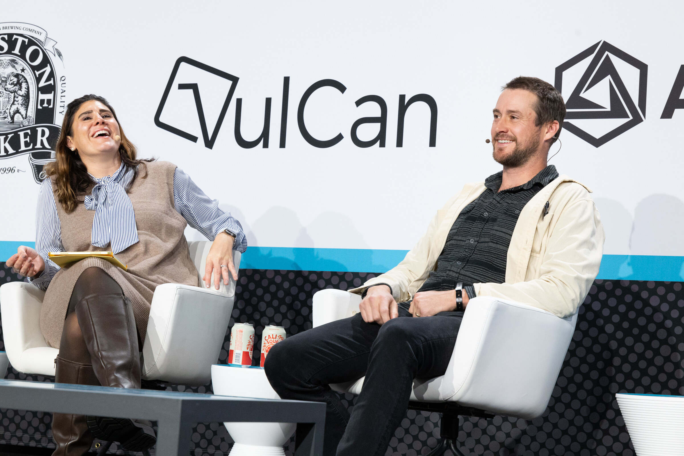 Woman in a tan vest laughing and a man in a cream jacket smiling on white chairs, with two red cans on the table between them.