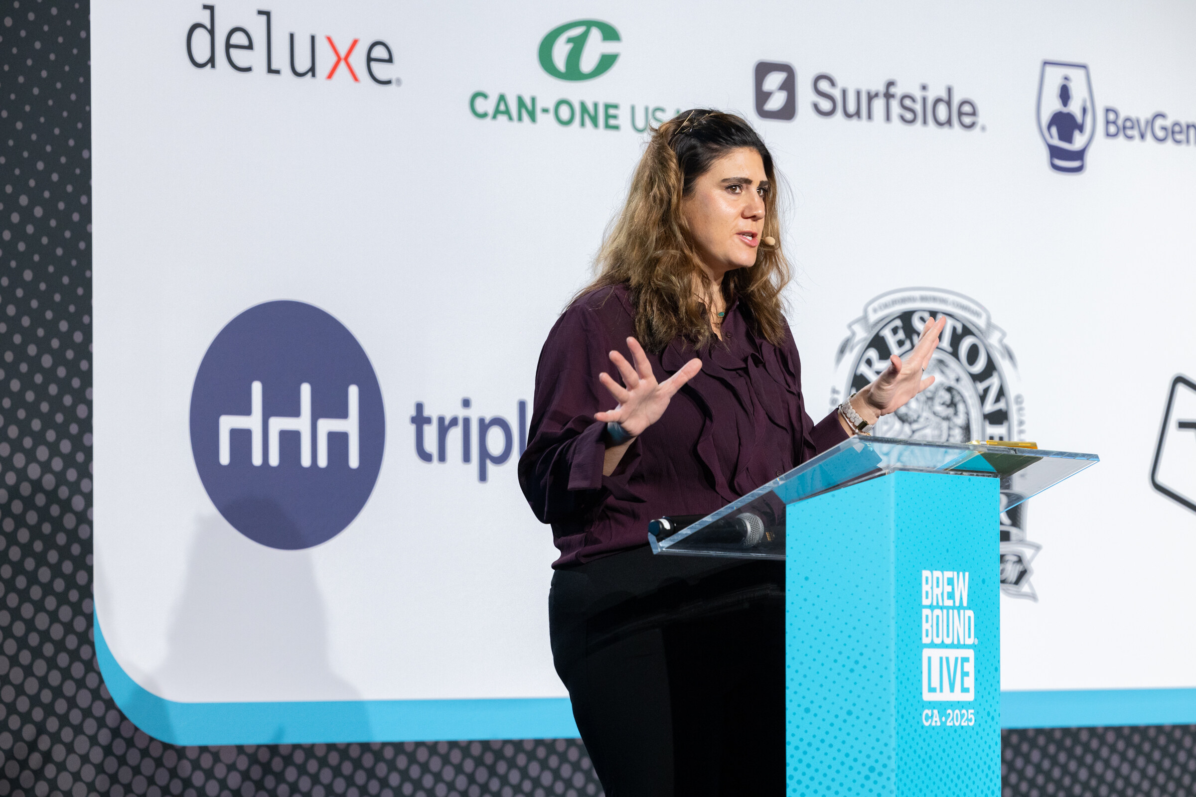 Woman in a plum blouse speaking at a teal Brewbound Live podium in front of Tripleseat and Firestone Walker logos.