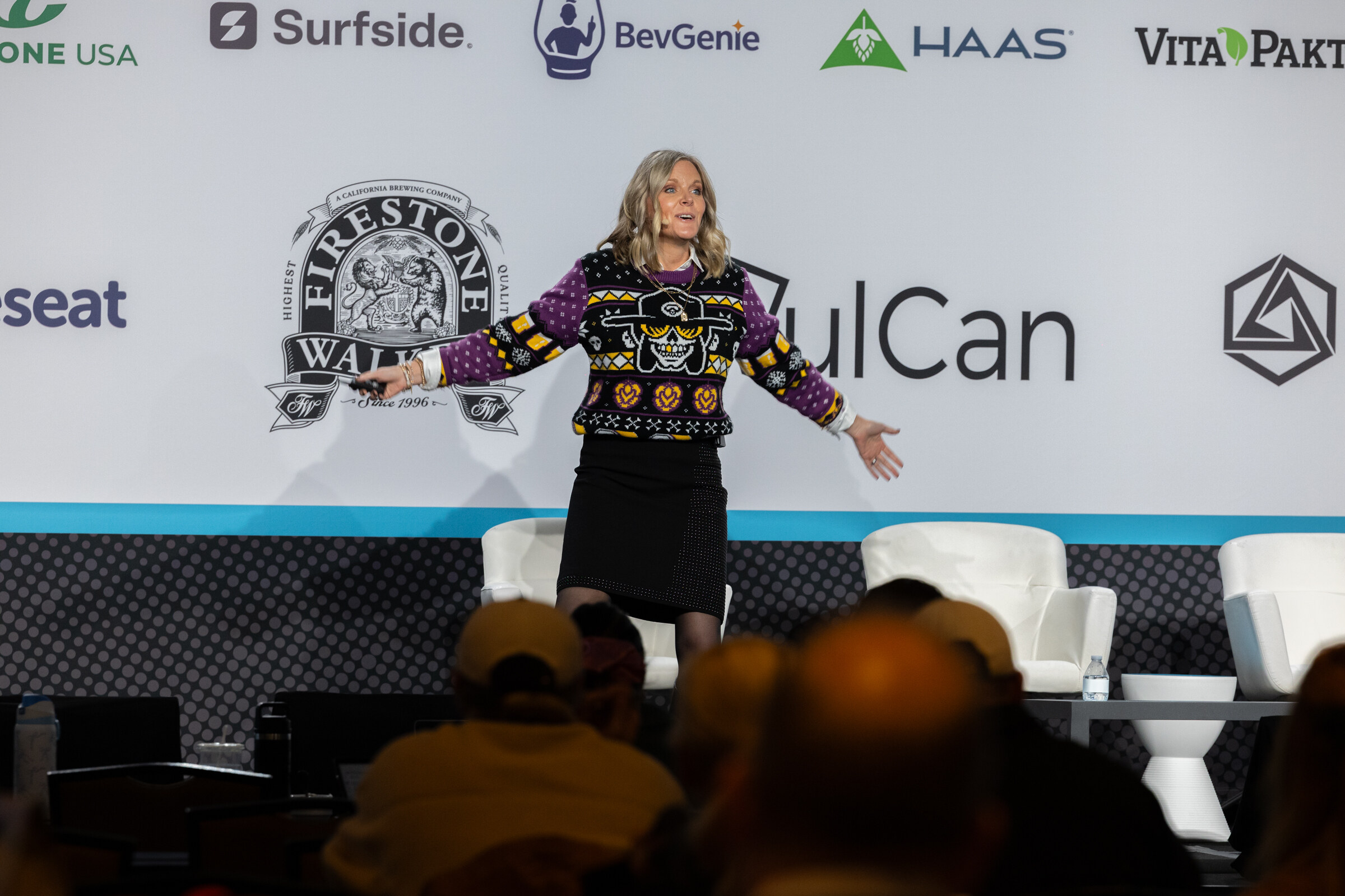 Woman in a colorful patterned sweater and black skirt standing center stage in front of a sponsor wall, with audience heads in the foreground.