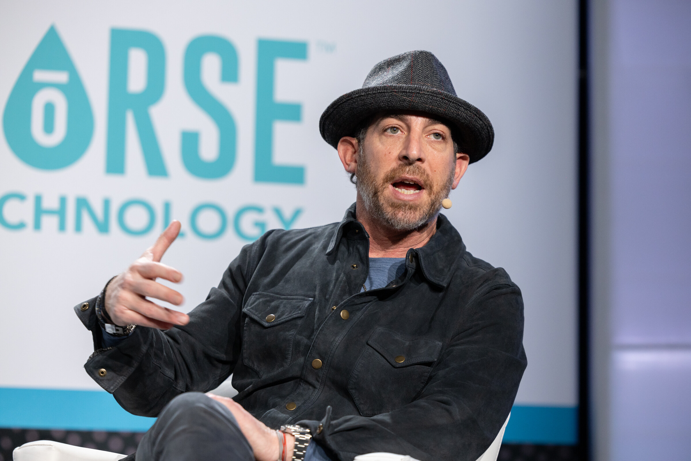 Man in a gray fedora and dark jacket gesturing while speaking in front of a Sorse Technology logo.