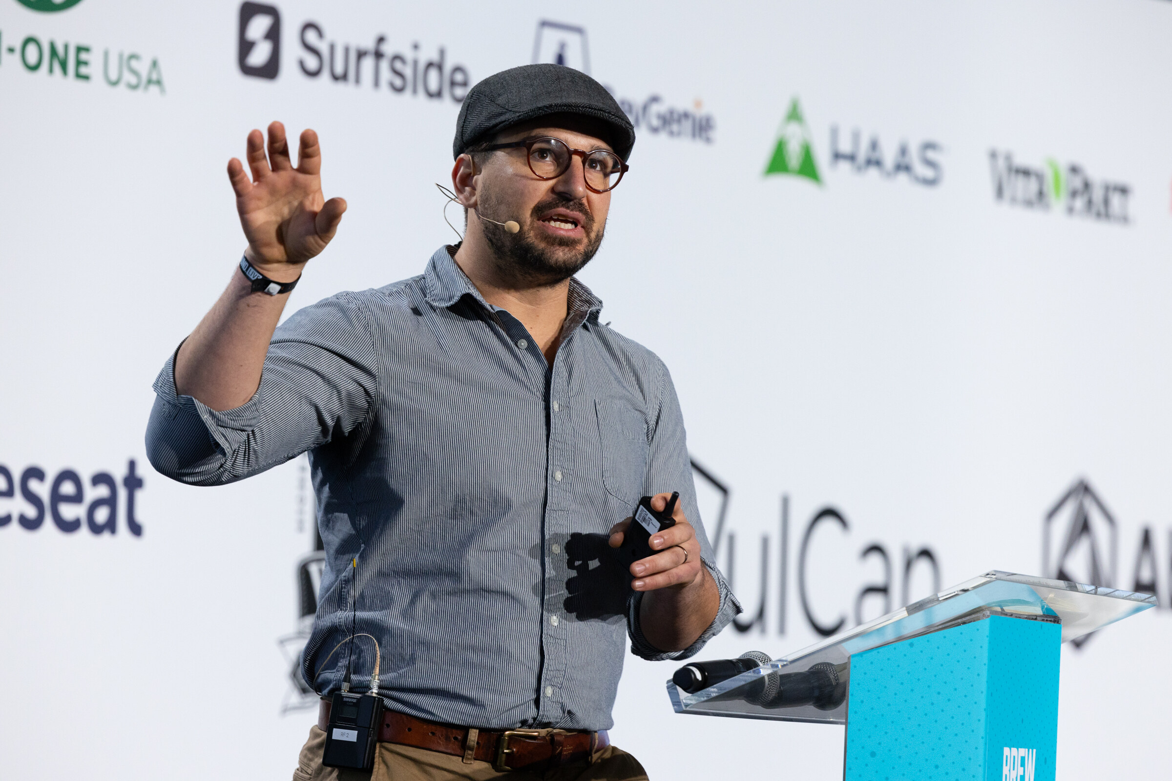 Man in a flat cap and glasses wearing a gray shirt and gesturing at a podium in front of a sponsor wall.
