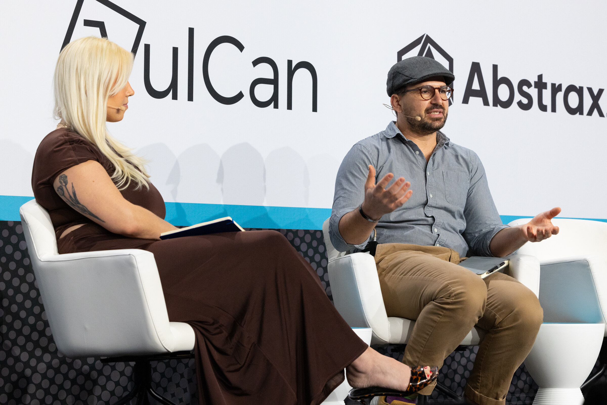Woman with long blond hair in a brown dress and a man in a flat cap seated on white chairs in front of VulCan and Abstrax logos.