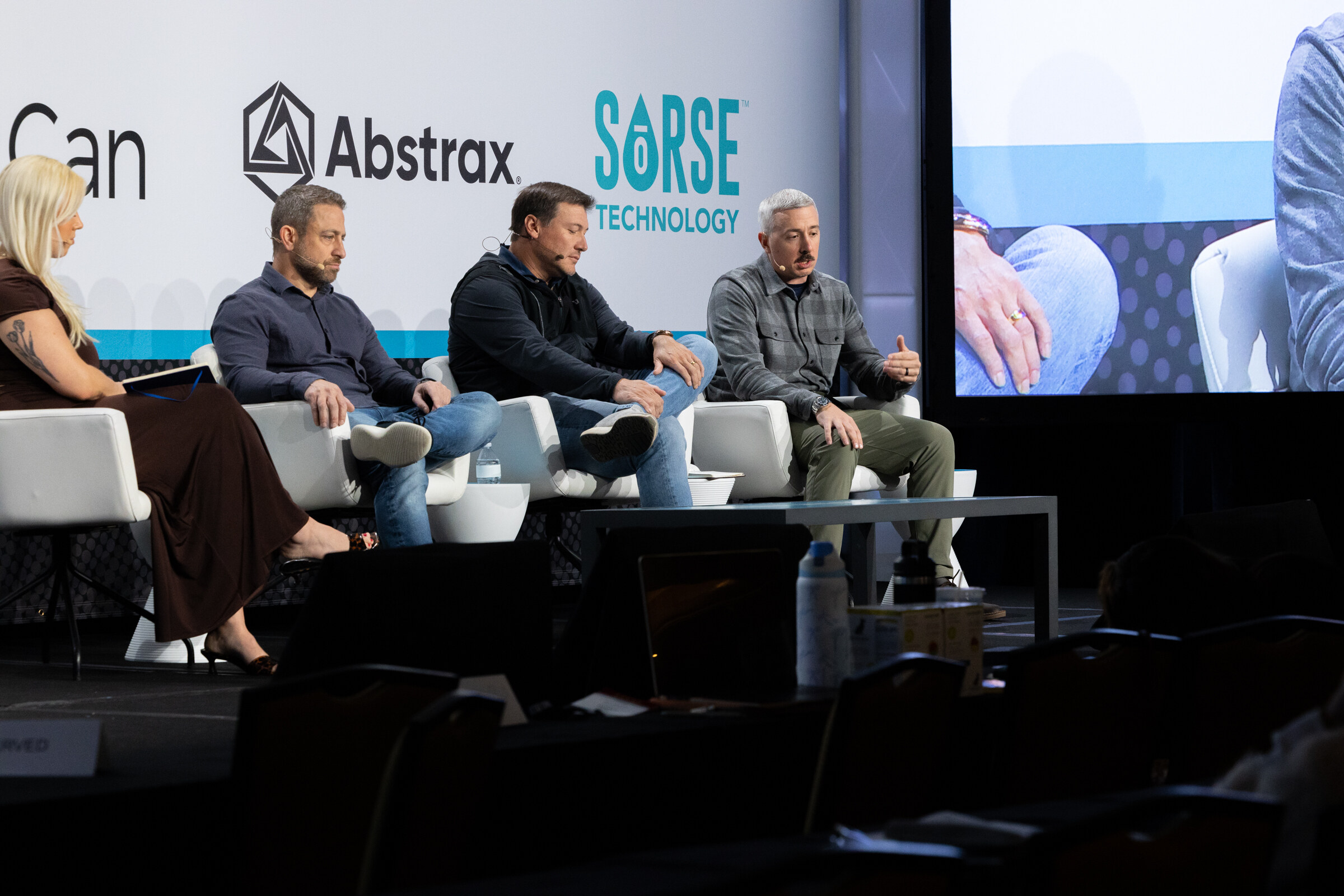Four people seated on white chairs on stage in front of VulCan, Abstrax and Sorse Technology logos with a video screen at right.