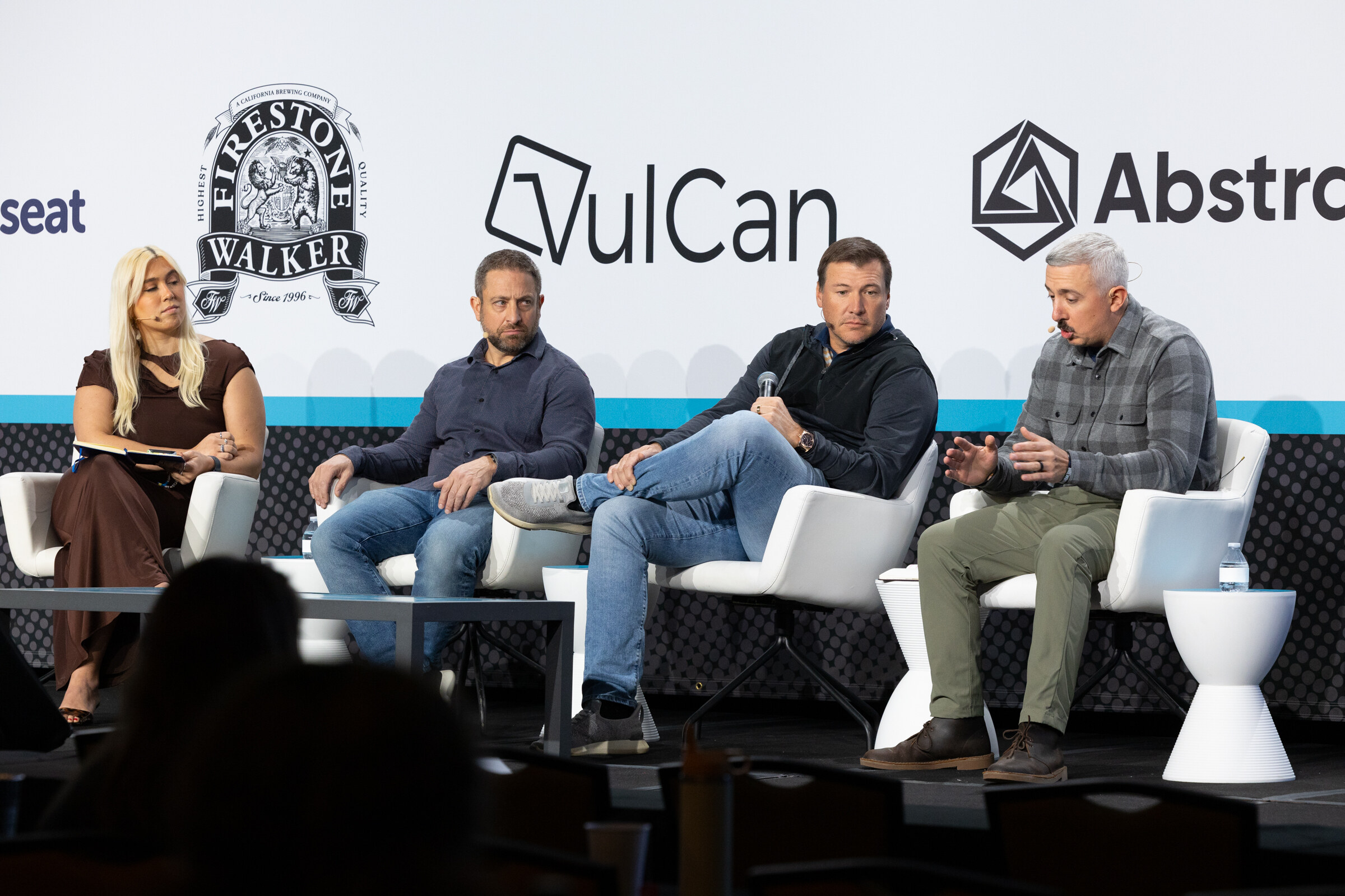 Woman with blond hair holding a notebook and three men seated on white chairs on stage in front of a sponsor wall.