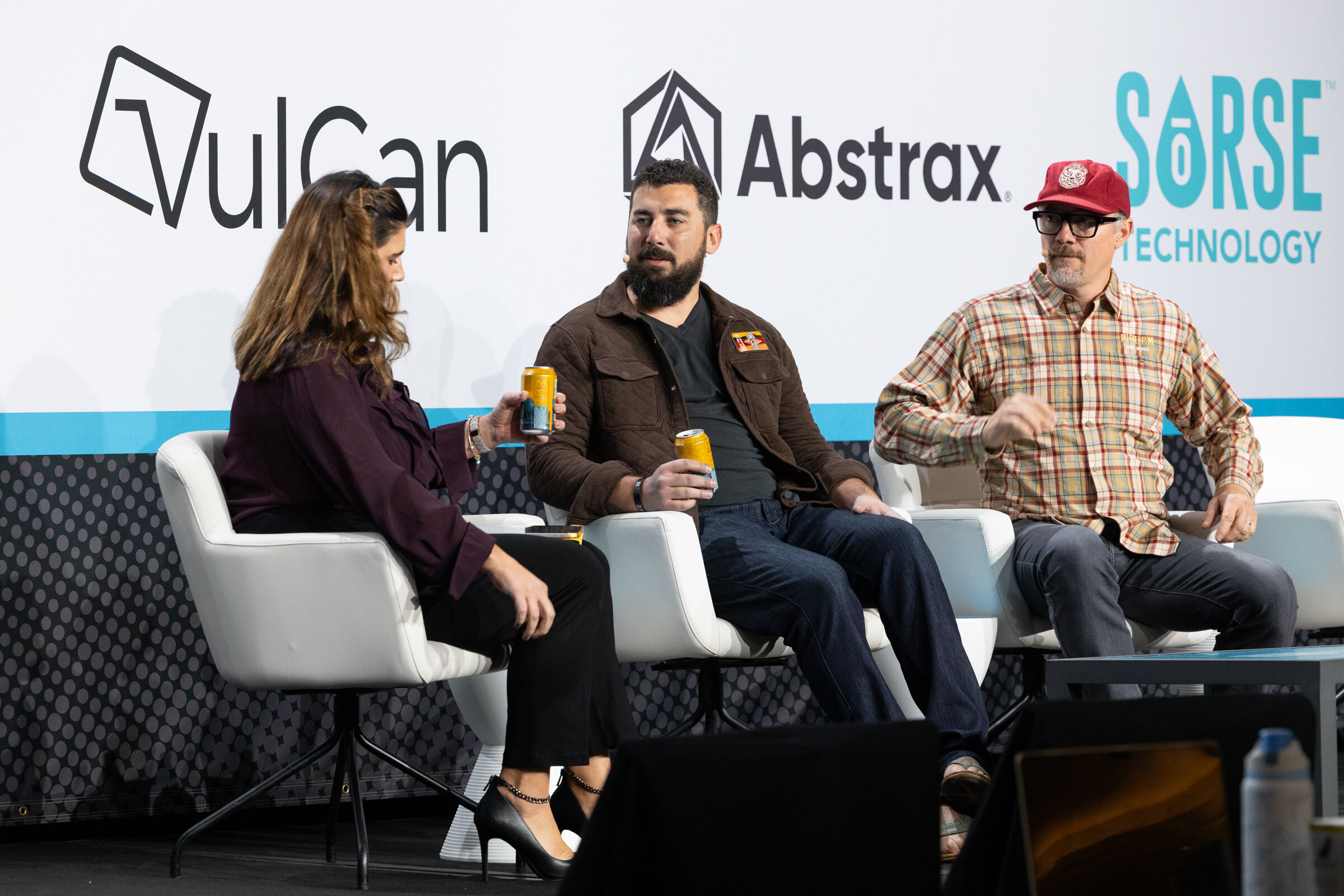 Woman in a plum top and two men holding yellow cans, seated on white chairs in front of VulCan, Abstrax and Sorse logos.