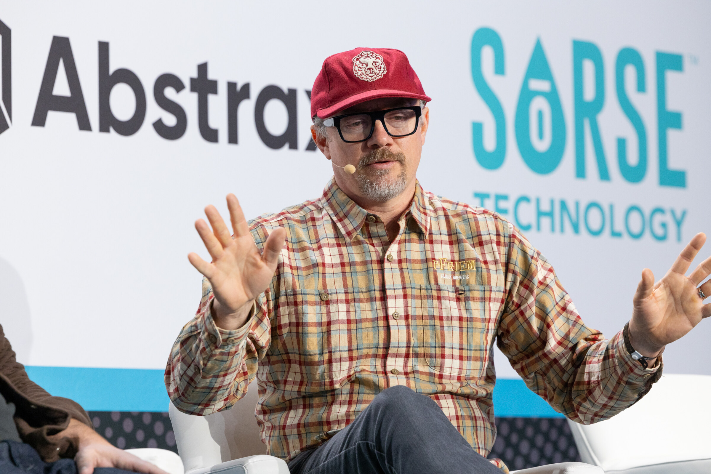 Man in a red cap, glasses and plaid shirt with both hands raised in front of Abstrax and Sorse Technology logos.