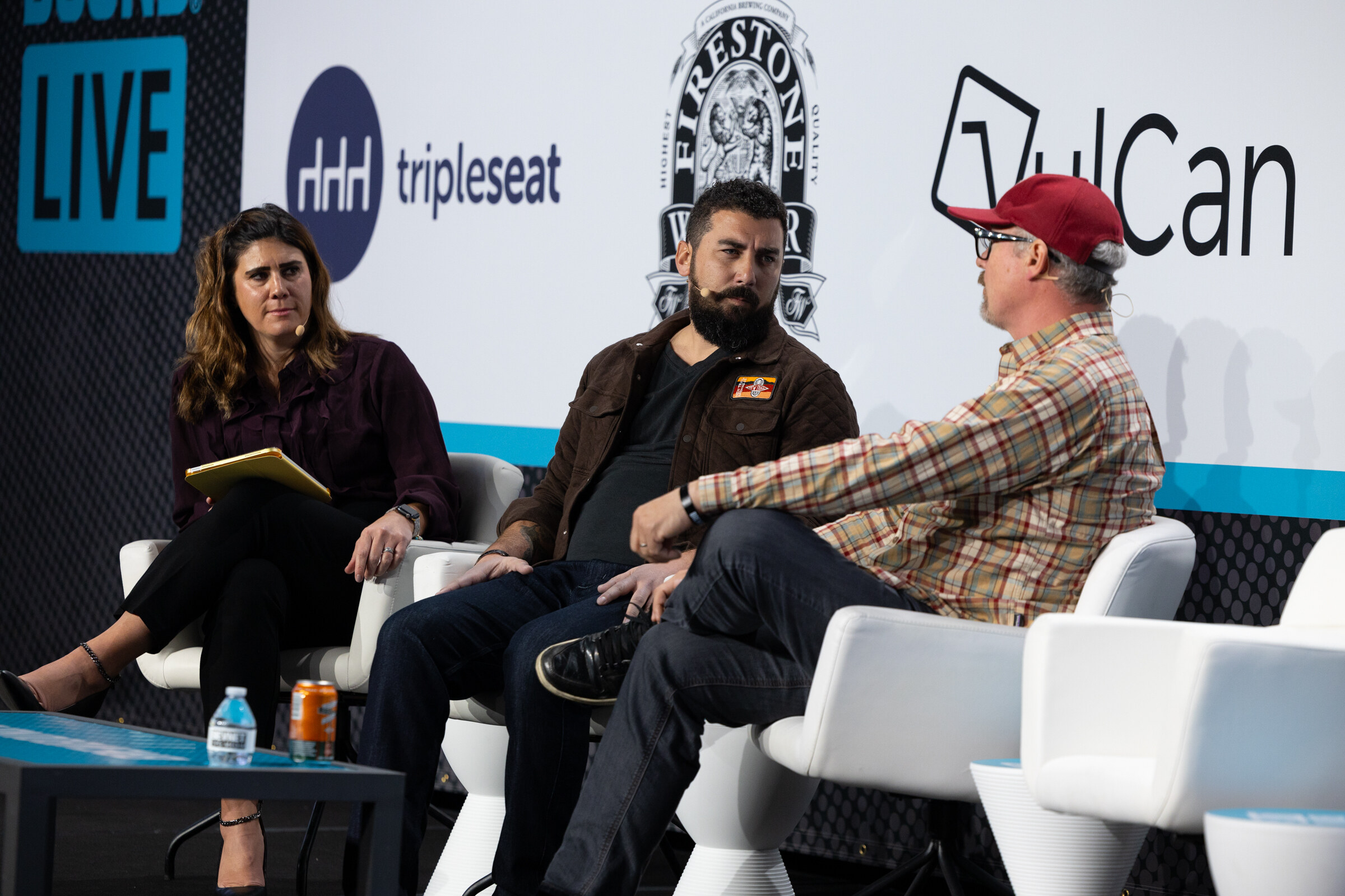 Woman with a tablet and two men on white chairs, with a water bottle and a can on the table, in front of Tripleseat and VulCan logos.