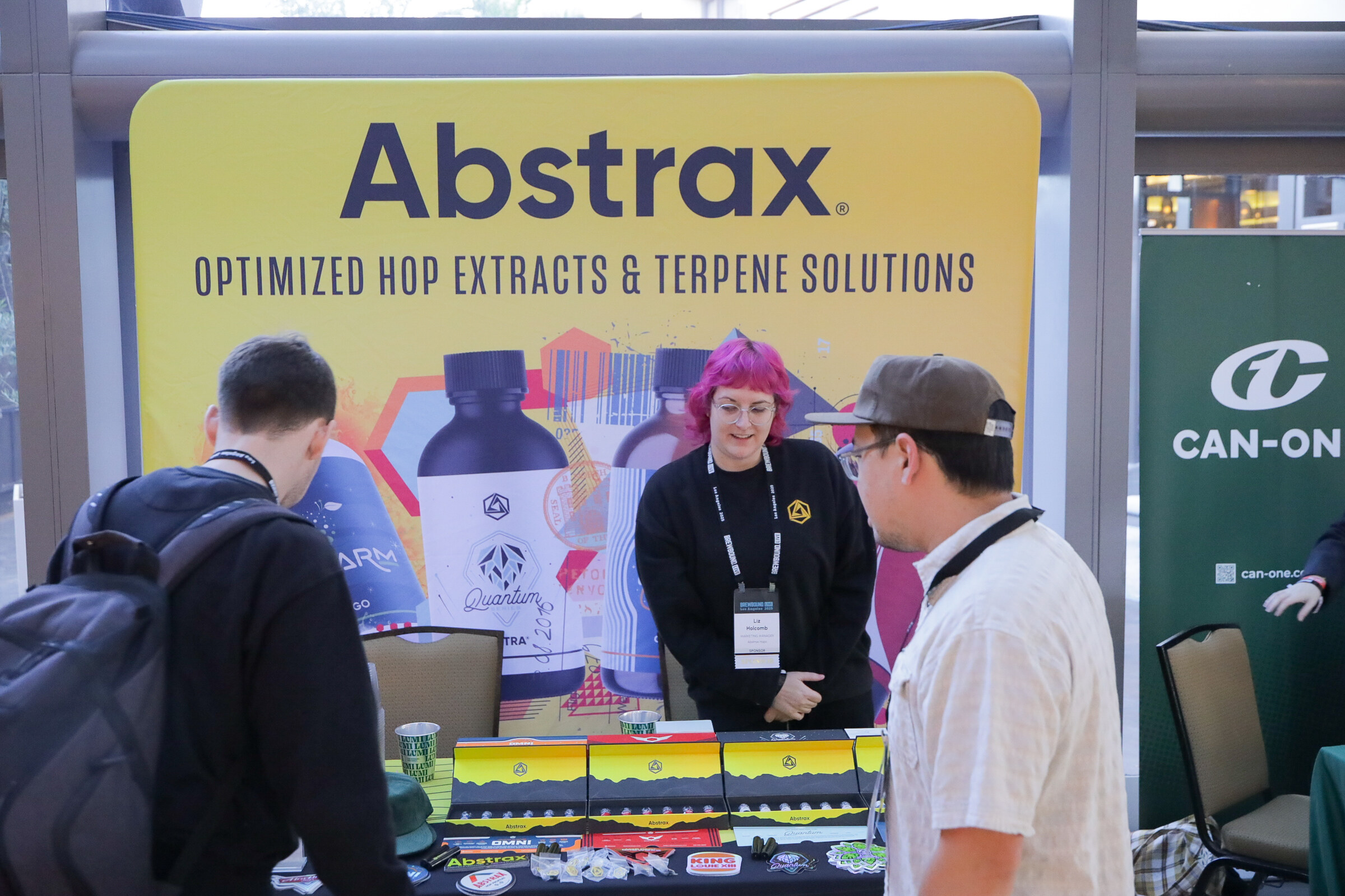 Woman with pink hair behind a table with product packaging in front of a yellow Abstrax banner, speaking with two attendees.