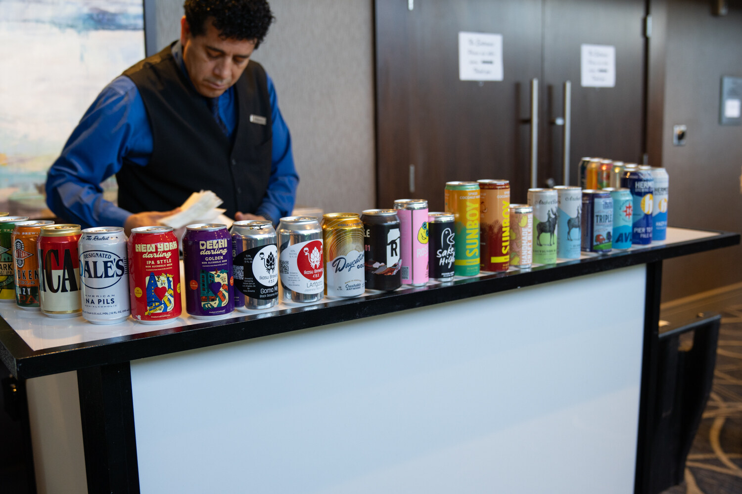 Man in a blue shirt and black vest arranging a long row of colorful cans on a white bar.