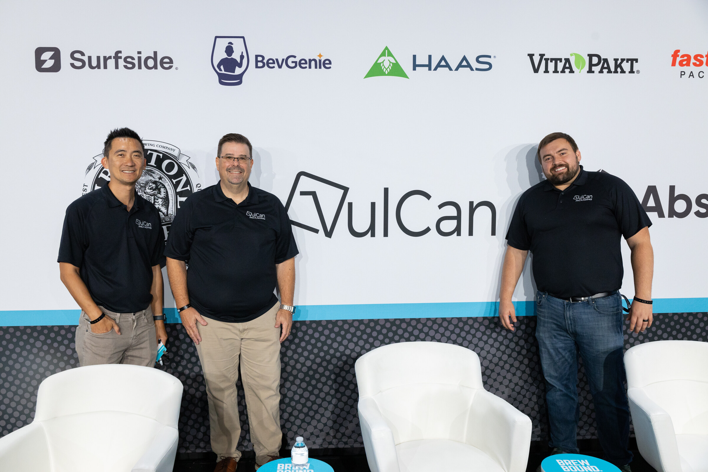Three men in black polo shirts standing in front of a sponsor wall with a VulCan logo and white chairs.