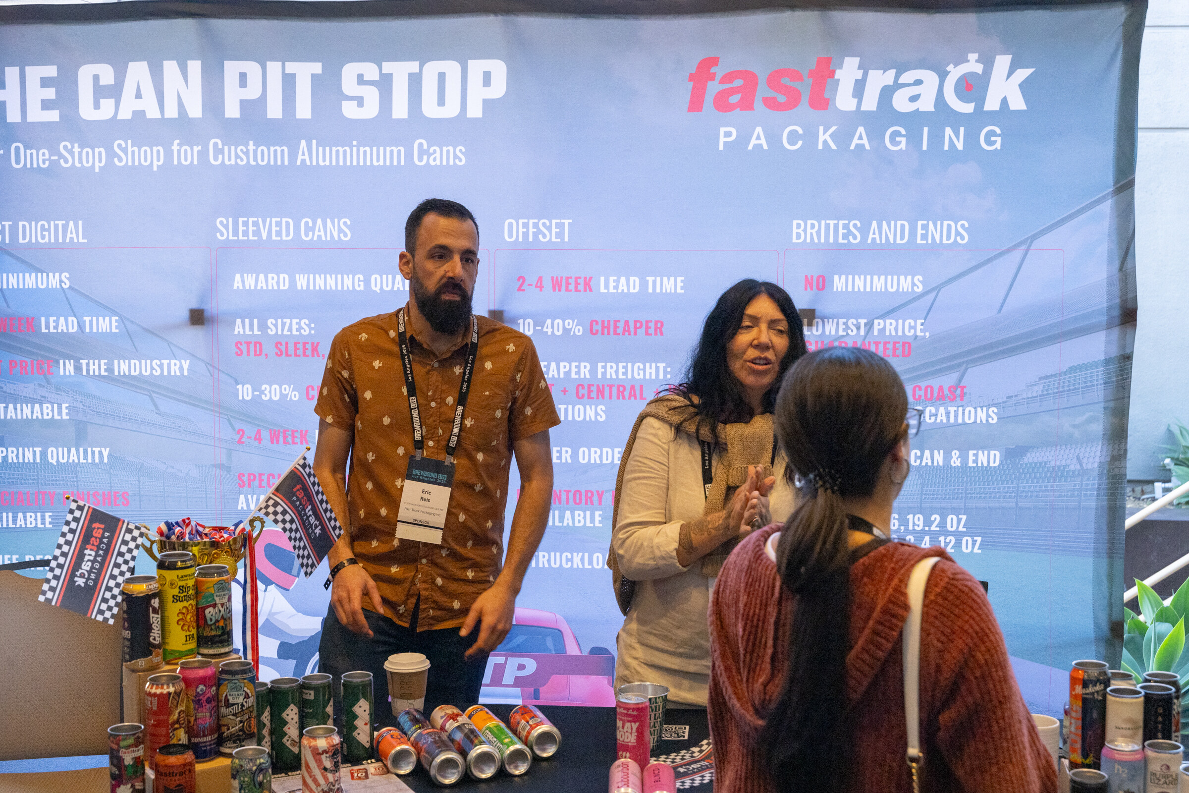 Man in a patterned shirt and a woman talking at a table of cans in front of a Fasttrack Packaging banner reading The Can Pit Stop.