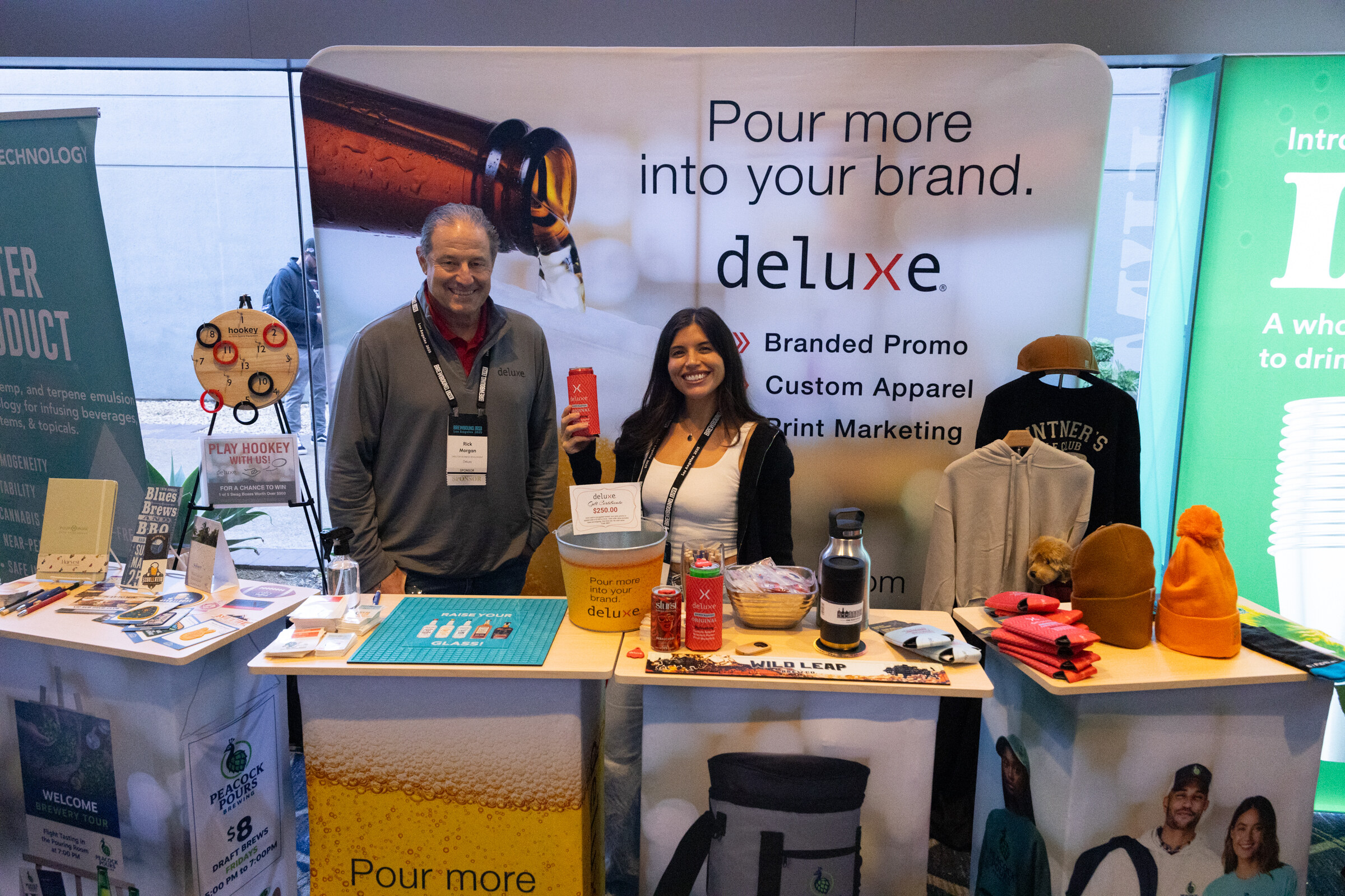 Man and woman with lanyards standing behind a table of branded apparel and drinkware in front of a Deluxe banner.