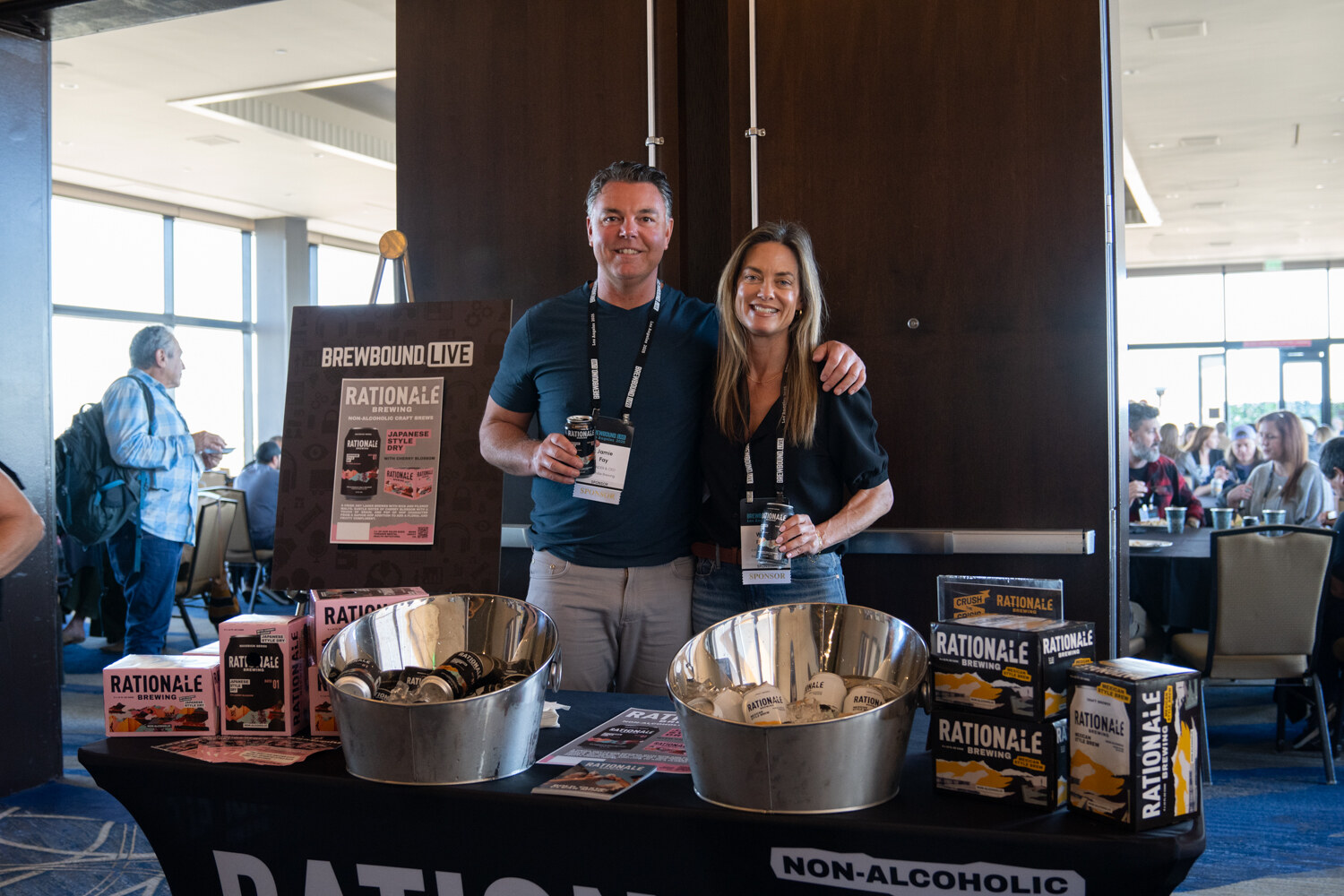 Man and woman with lanyards holding cans behind a table with a Rationale Brewing tablecloth, buckets of cans and boxes of non-alcoholic beer.
