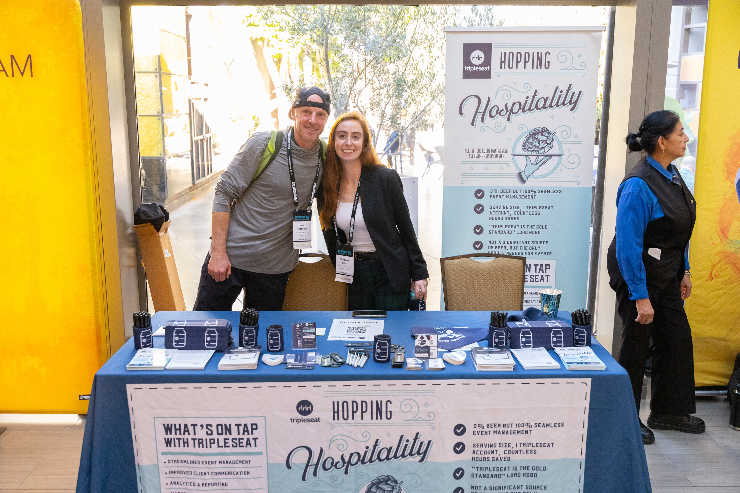 Man in a cap and woman in a black blazer standing behind a blue table with Tripleseat banners and giveaways.