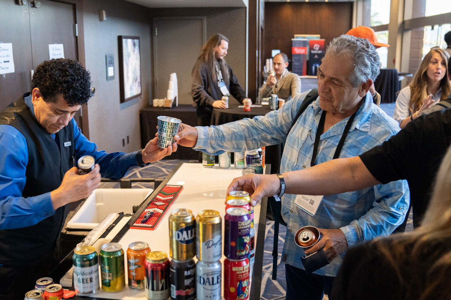 Man in a blue shirt and vest pouring a sample while a man in a light blue shirt holds out a cup beside a bar covered in cans.