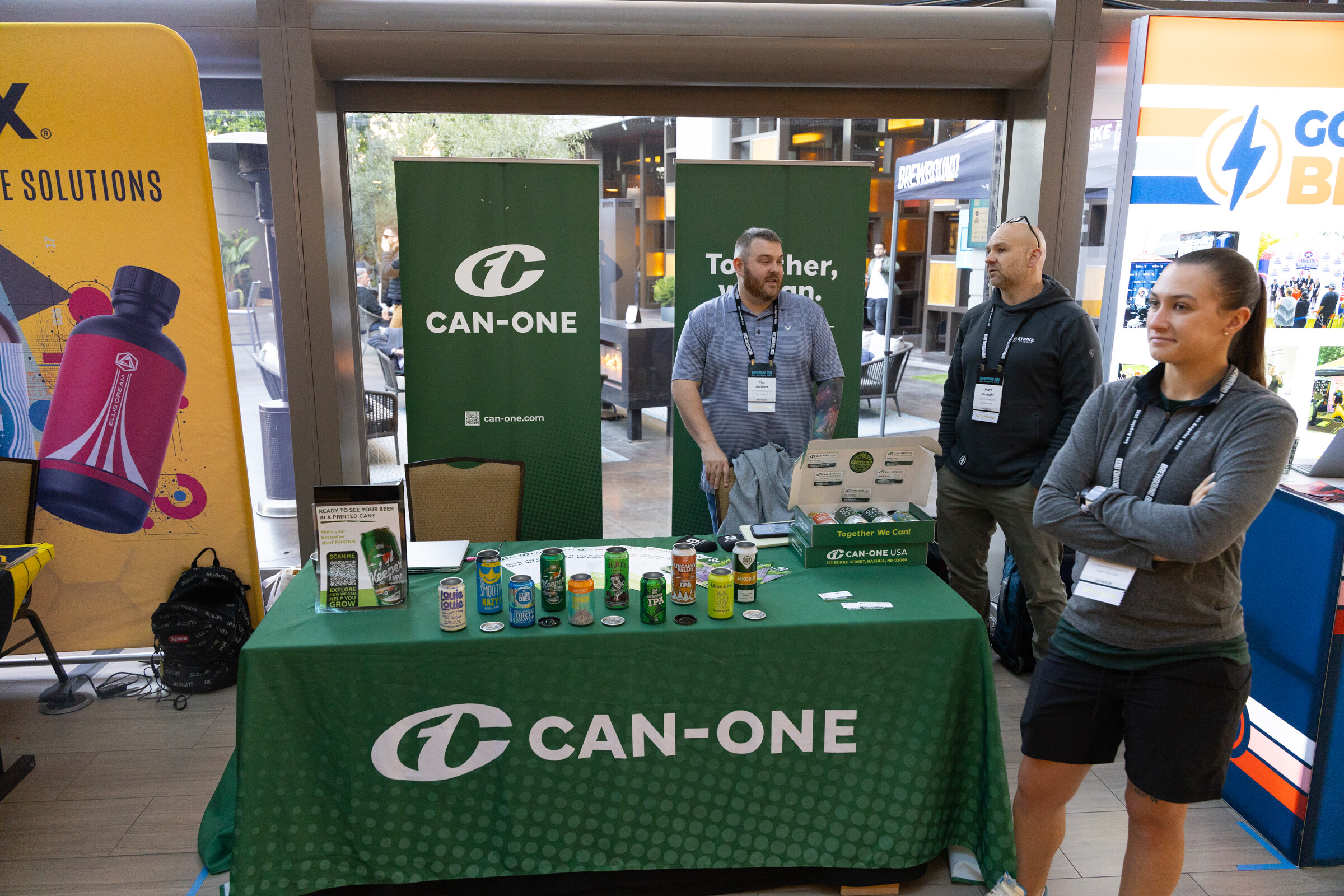 Three people behind a green Can-One table with cans, in front of Can-One banners.