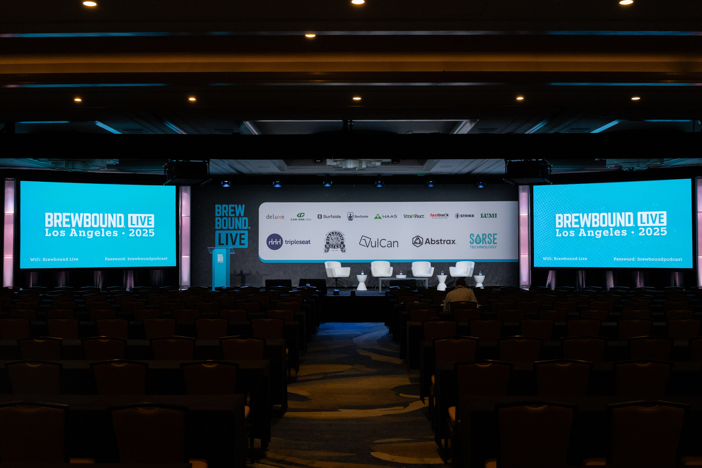 Dark ballroom with rows of chairs facing a stage, two screens reading Brewbound Live Los Angeles 2025, and a sponsor wall.
