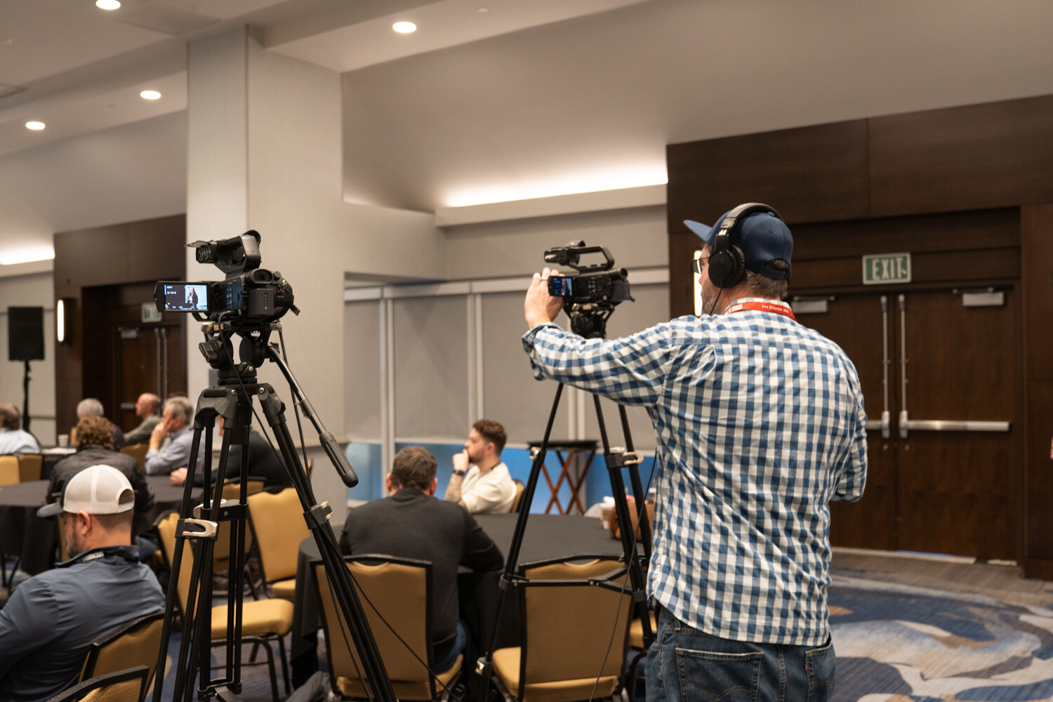 Man in a checked shirt and headphones filming with a camera on a tripod in a meeting room.