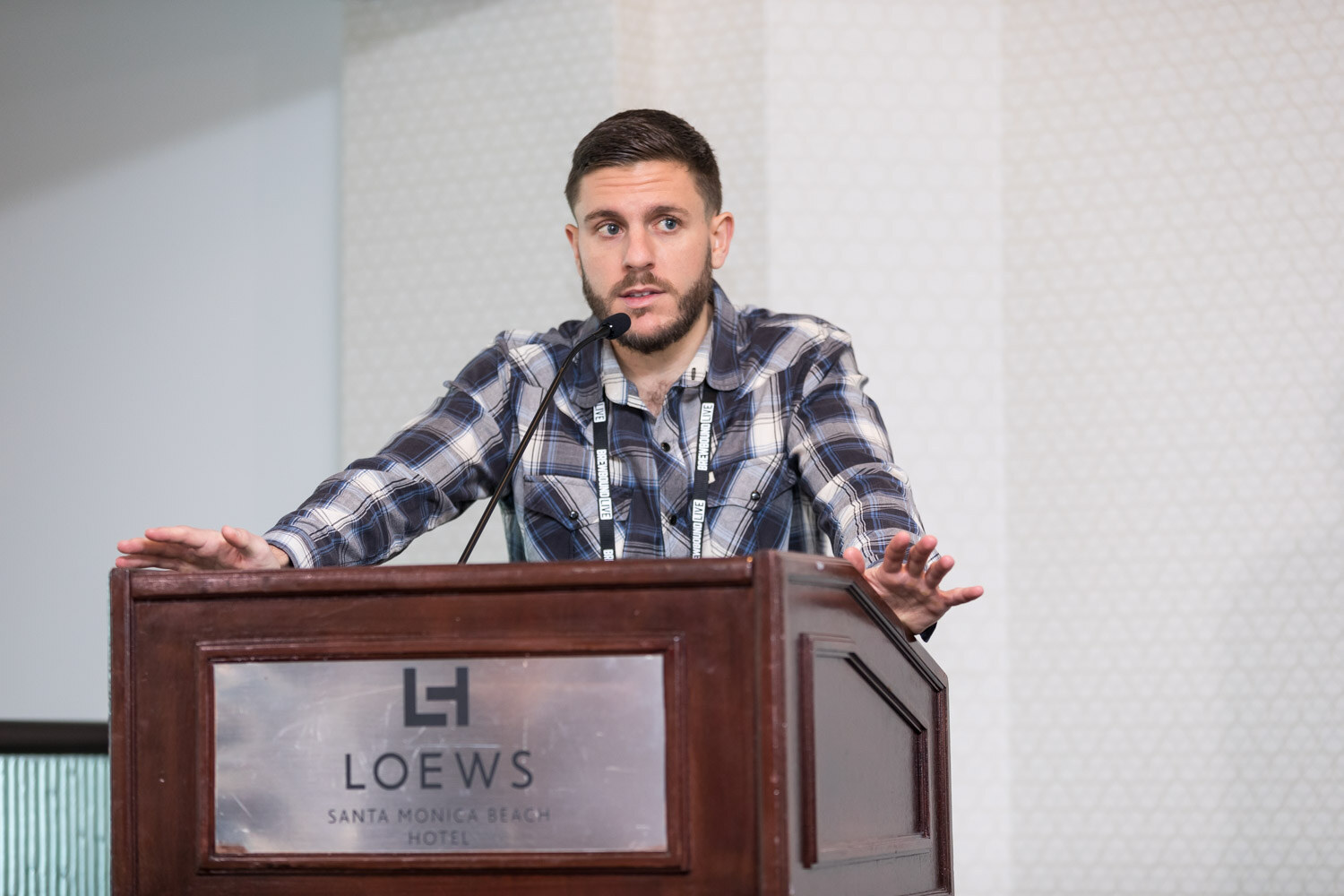 A bearded man in a navy plaid shirt rests his hands on a wooden podium with a Loews Santa Monica Beach Hotel plate.