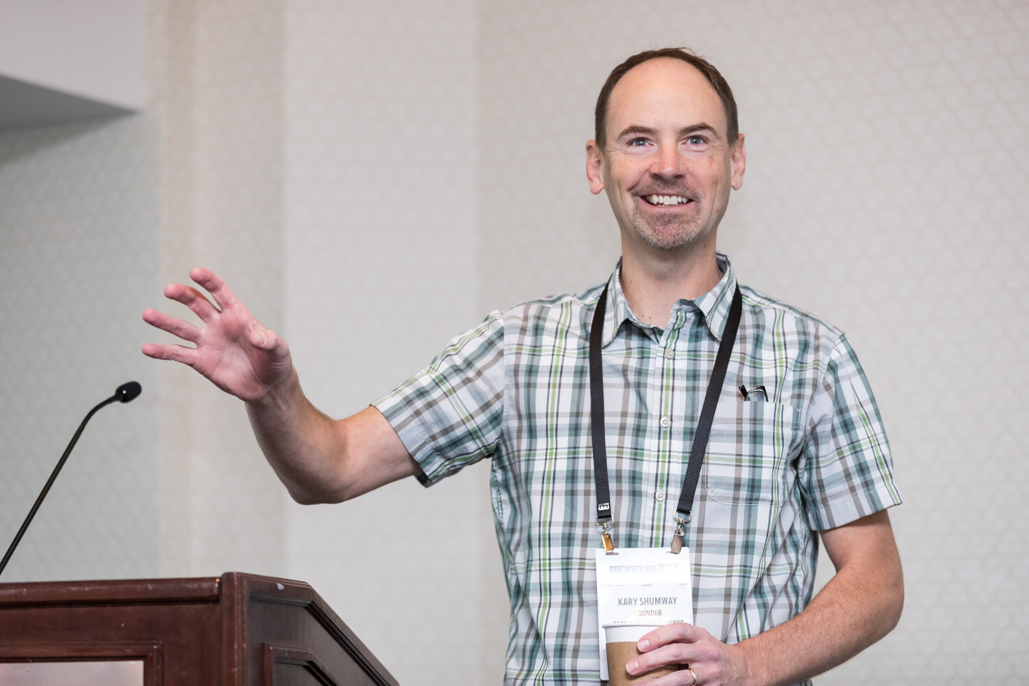 A man in a plaid short-sleeve shirt and lanyard gestures beside a lectern in a hotel meeting room.