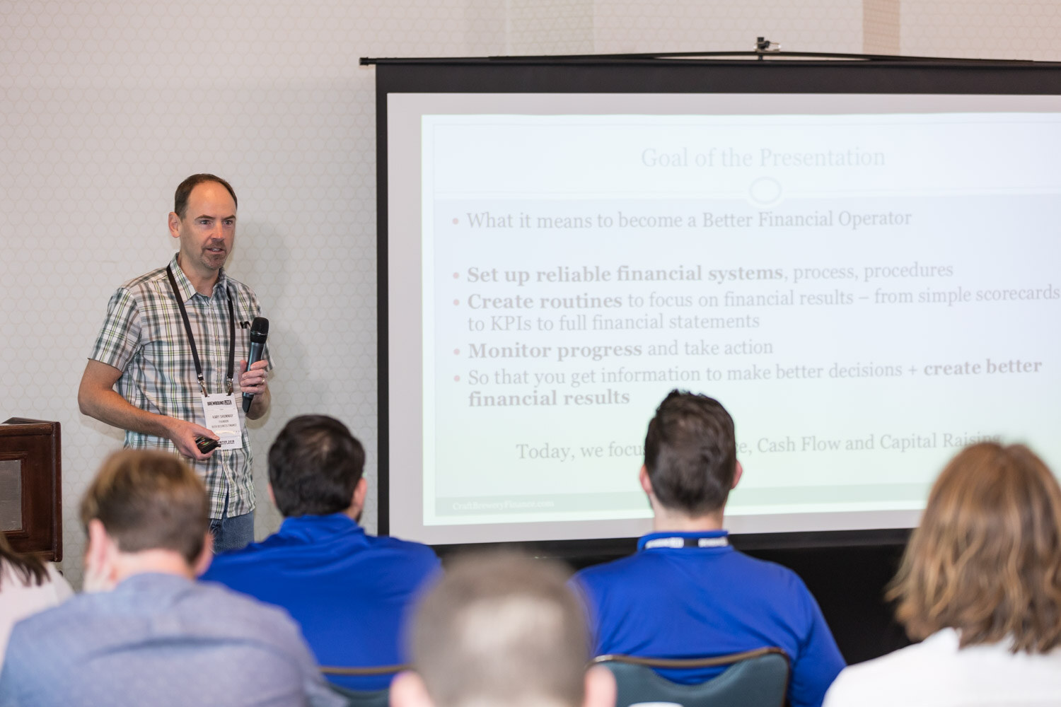 A presenter holding a microphone stands beside a projection screen of bullet points about financial systems, with attendees seen from behind.