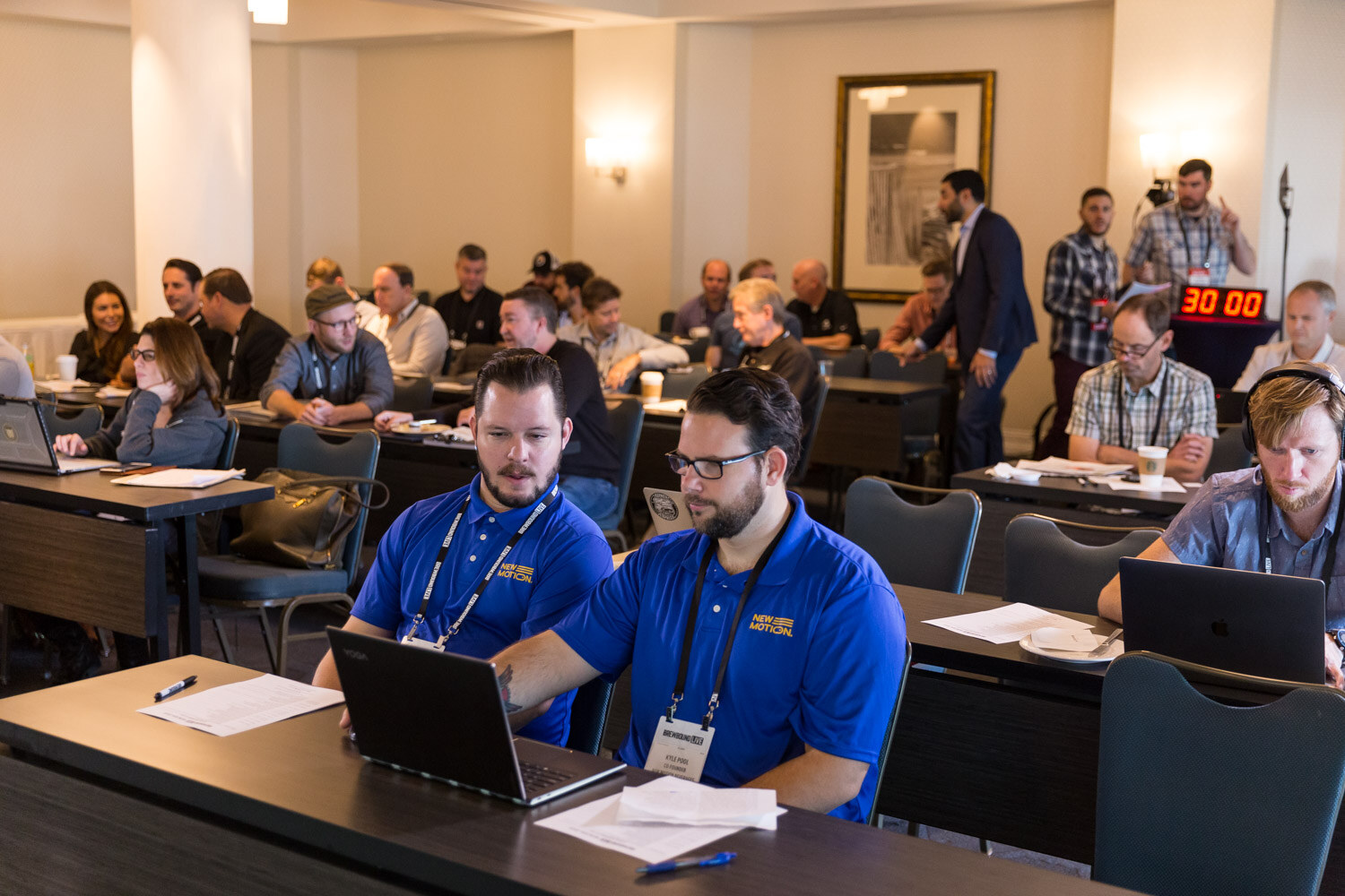 Rows of attendees at long tables, two men in royal blue polo shirts working on laptops in the foreground and a countdown timer in the background.