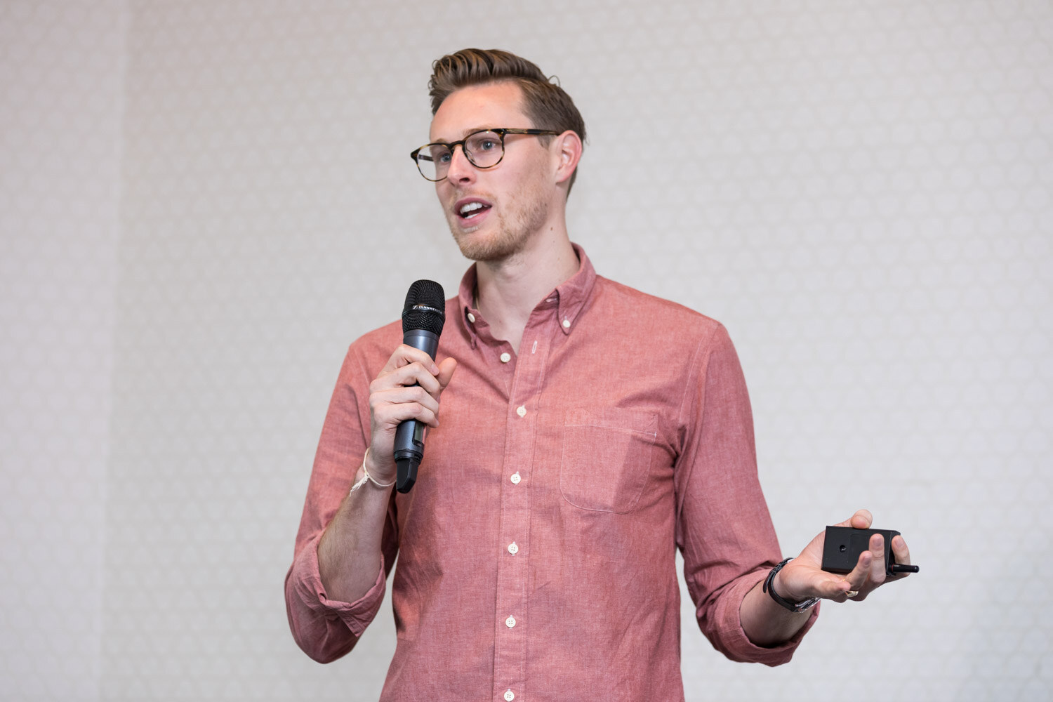 A man with glasses and a coral shirt holds a microphone and a clicker in a hotel meeting room.