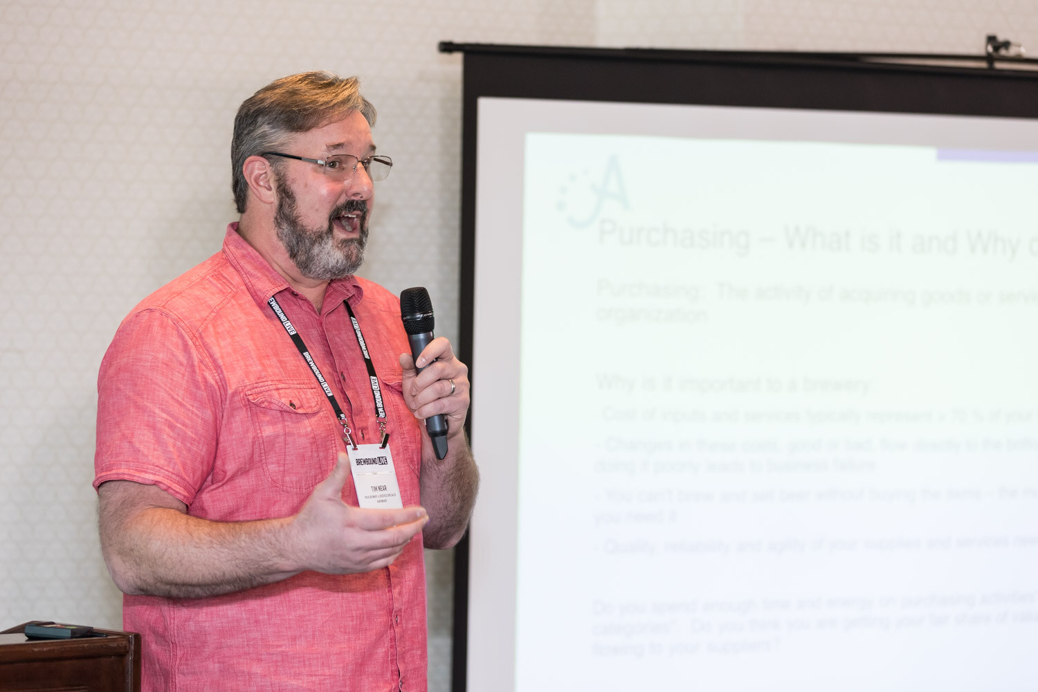 A bearded man with glasses and a lanyard speaks into a microphone next to a projection screen with a Purchasing slide.
