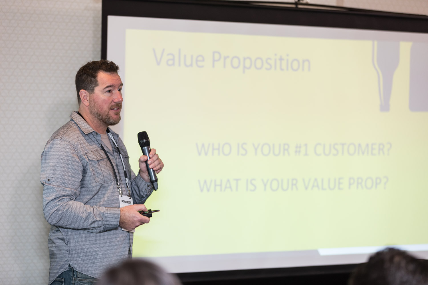 A man in a gray button-up shirt holds a microphone beside a yellow-green slide with the text Who is your number one customer? What is your value prop?