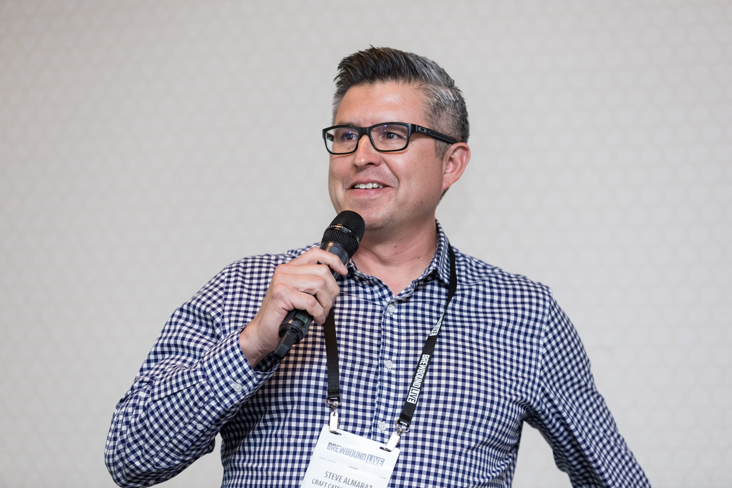A smiling man with glasses and a lanyard holds a microphone against a plain wall.
