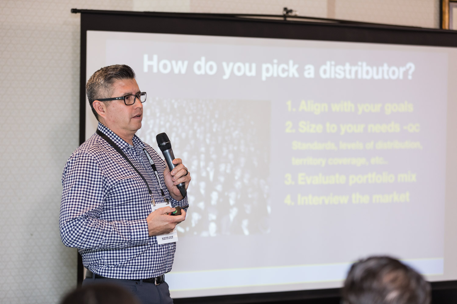 A man with a microphone stands at the edge of a projection screen showing a numbered list under the heading How do you pick a distributor?