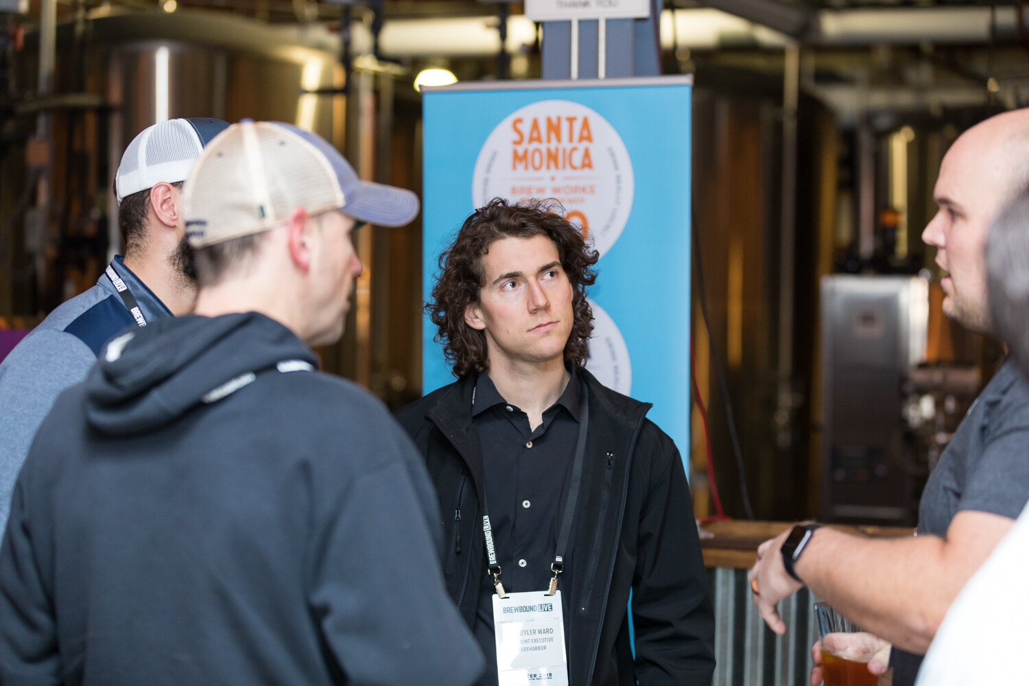 A young man with curly hair in a black jacket stands near stainless tanks and a Santa Monica Brew Works banner while others talk.