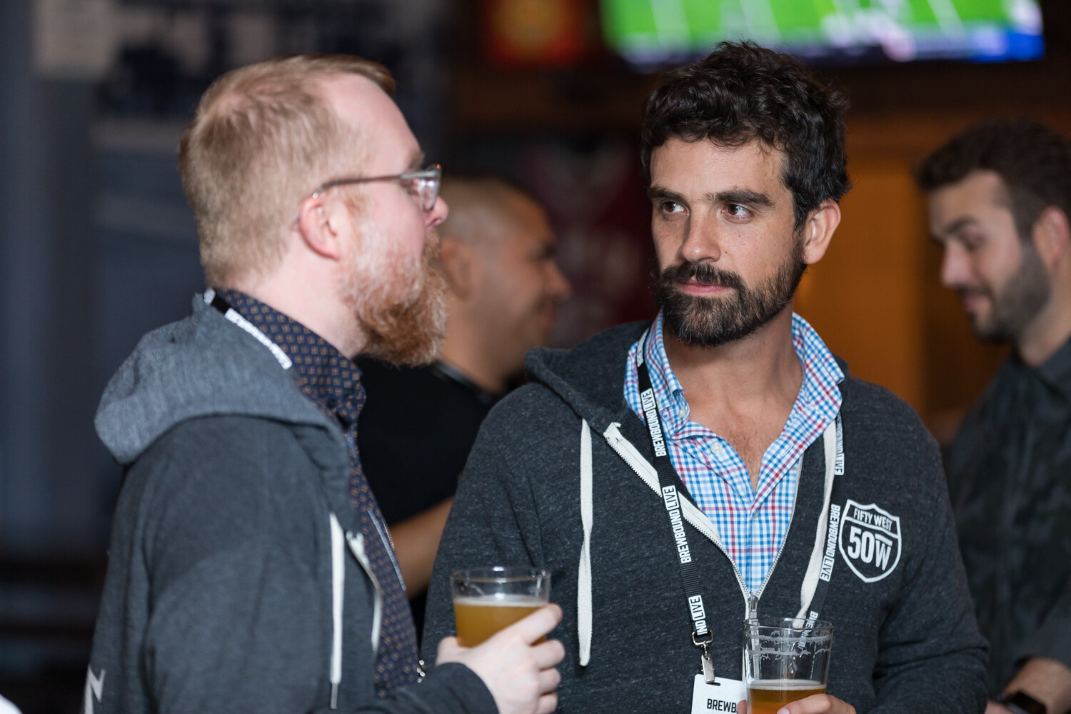 Two men with lanyards and beer glasses talk in a dim taproom.