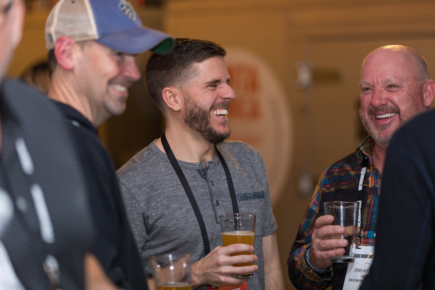 Three men with lanyards and glasses of beer laugh together in a warmly lit taproom.