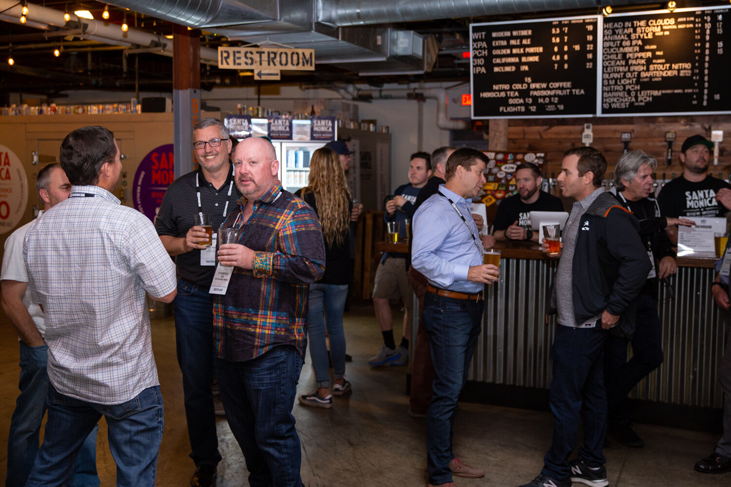A crowded taproom with attendees holding beers near a bar with a chalkboard tap list.
